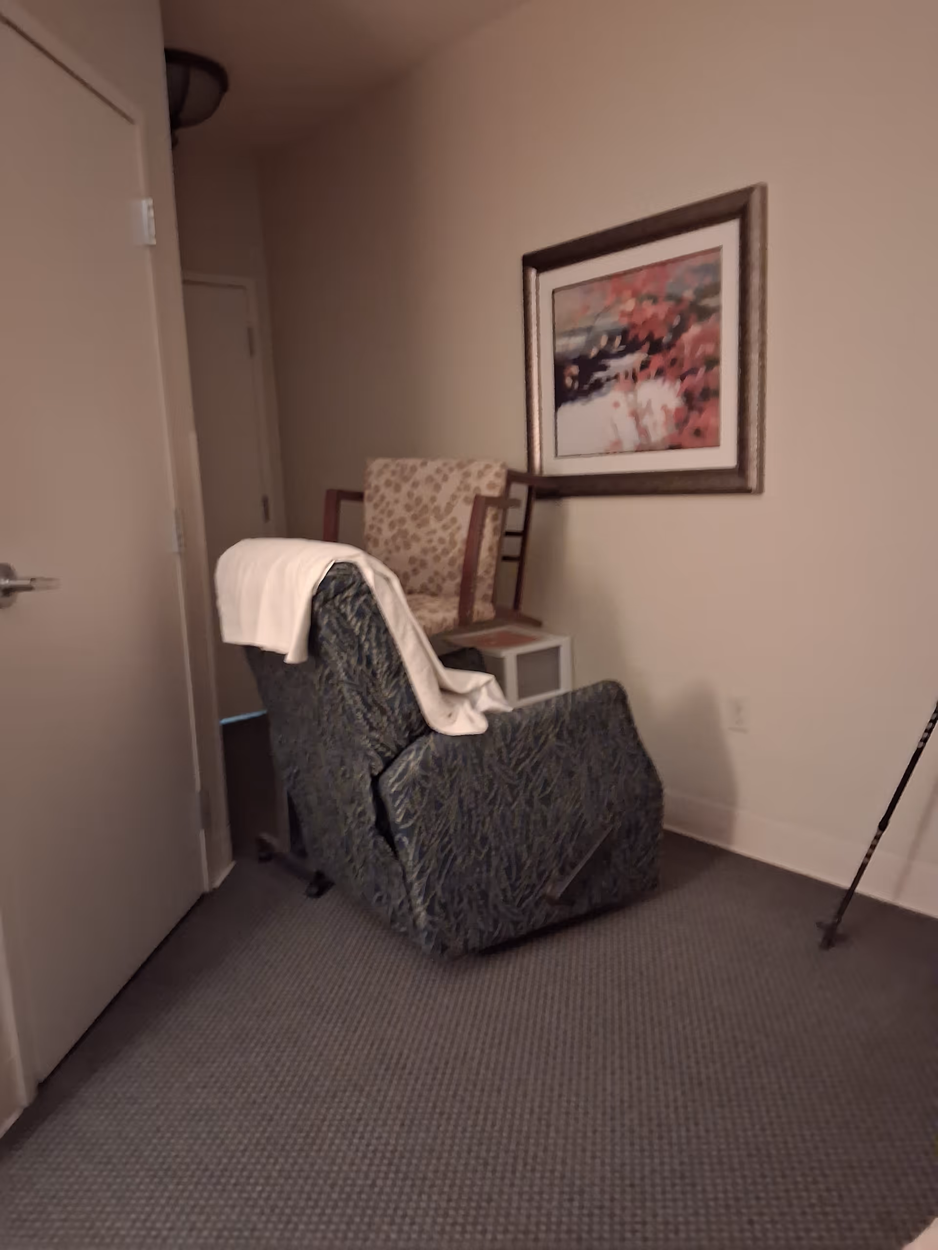 Interior of a senior living facility room with a recliner, patterned chair, and wall art.