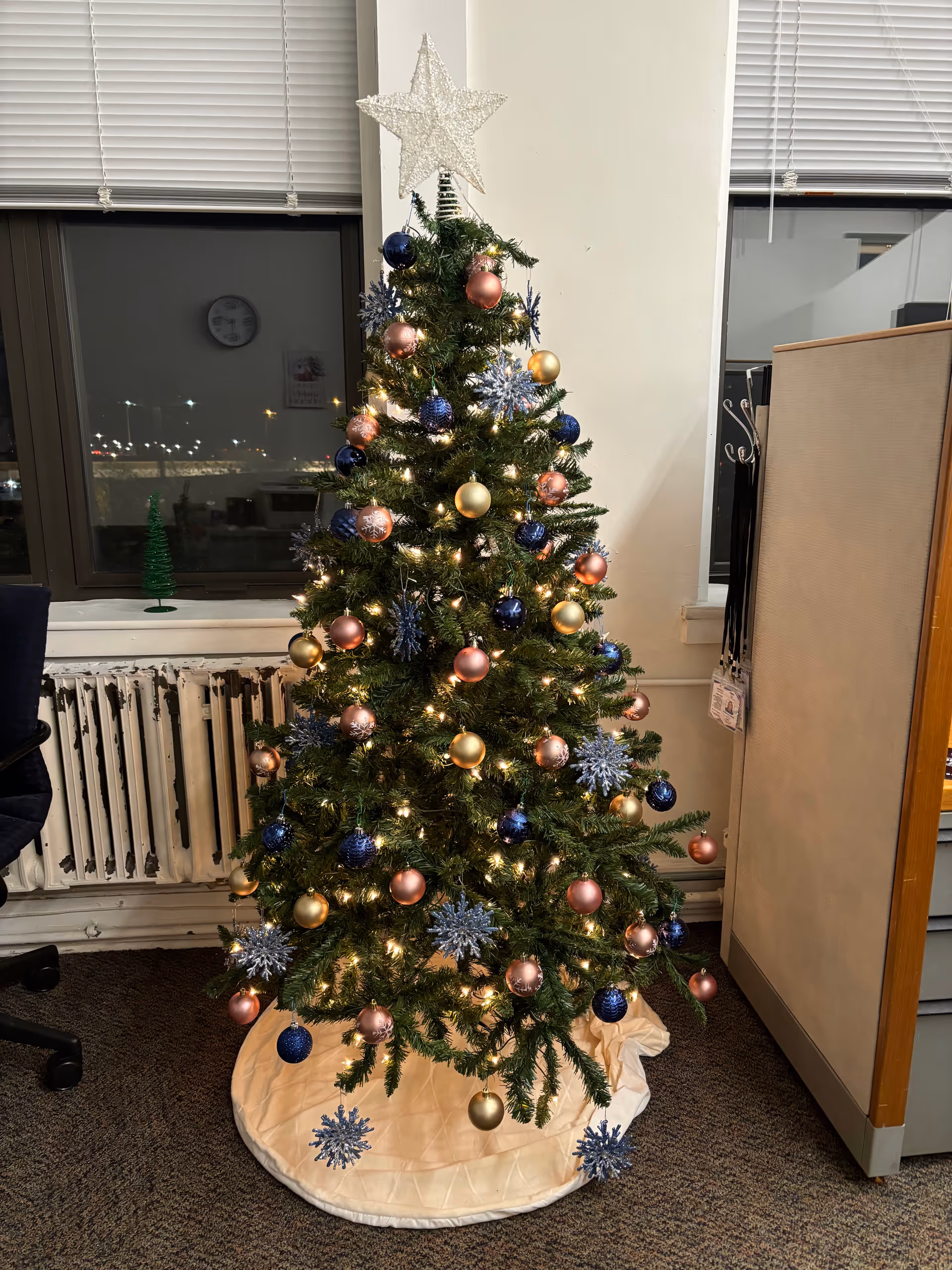 An indoor Christmas tree with lights and colorful ornaments stands beside a radiator in an office.