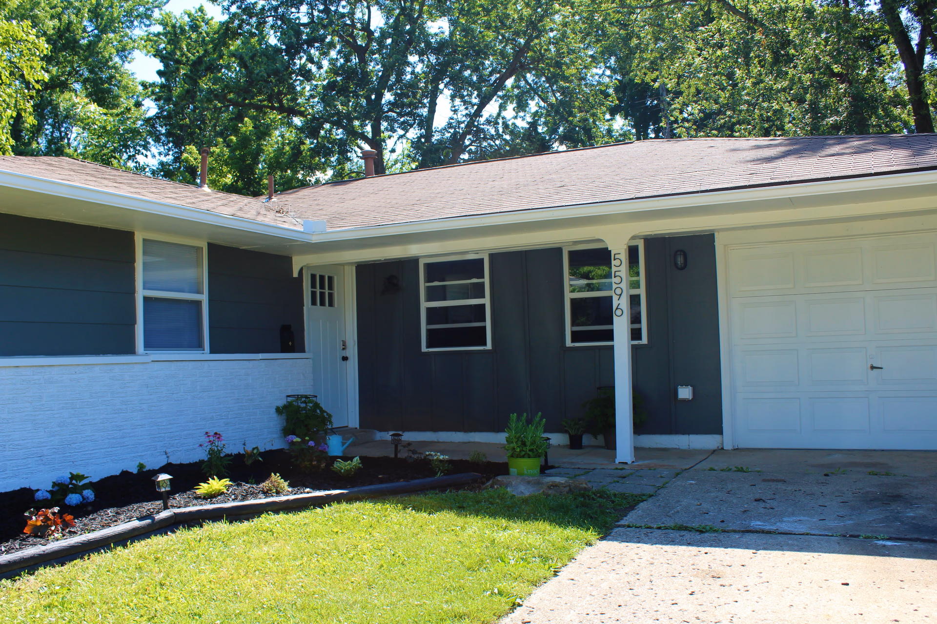 Front exterior view of a single-story house with a white door, two windows, a garage door, and a small garden bed with plants and flowers. The house has dark gray siding with white trim and a white pillar with the house number 5596. Trees are visible in the background.