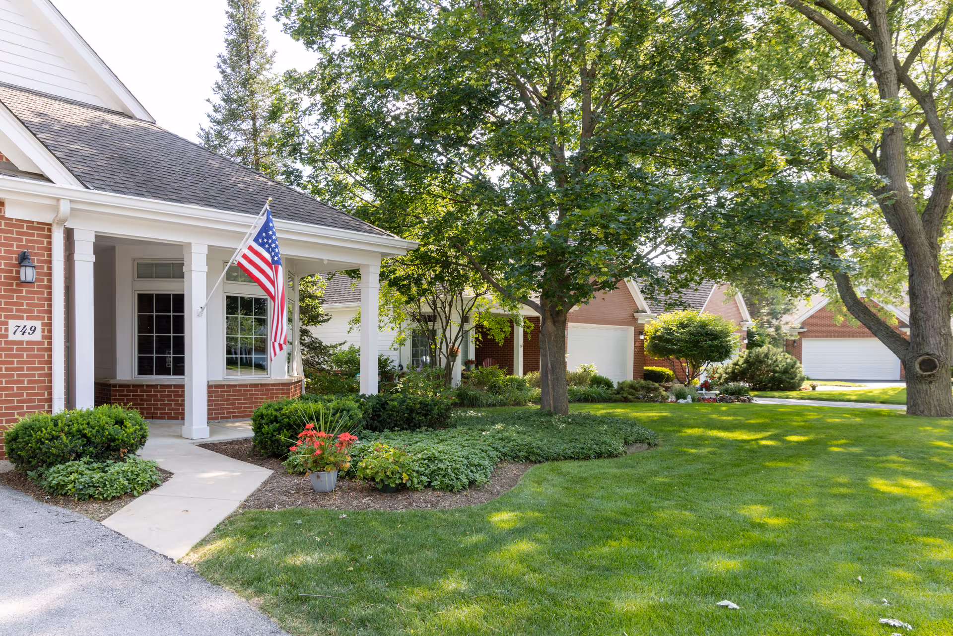 Front exterior of a brick house with a covered porch, American flag, and landscaped lawn with trees.