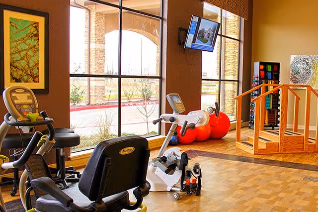Bright rehabilitation exercise room featuring recumbent bikes, pedal machines, large red therapy balls, a set of steps and a wall-mounted TV in front of large windows.