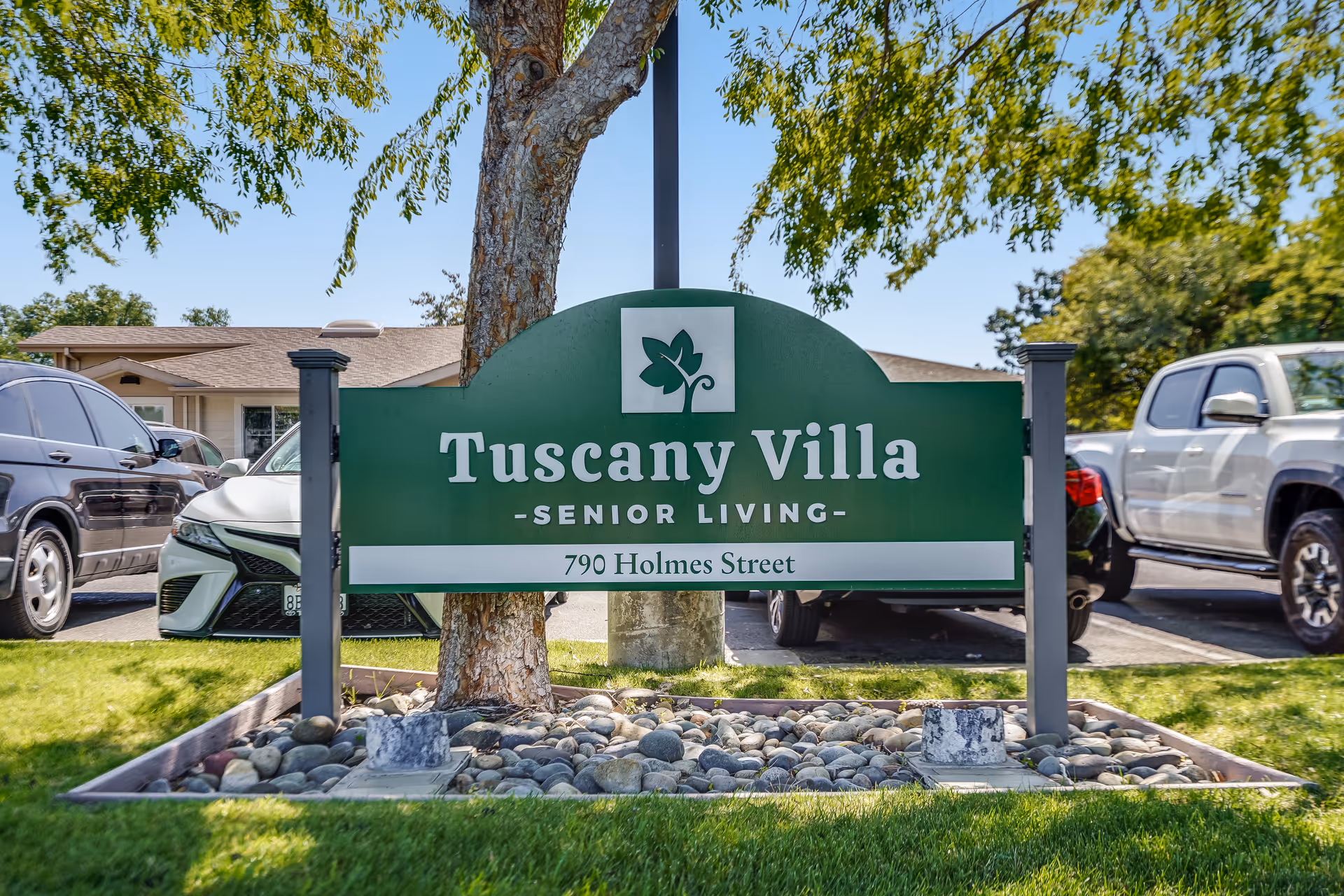 Green entrance sign reading "Tuscany Villa Senior Living - 790 Holmes Street" mounted in front of a tree with parked cars behind it.