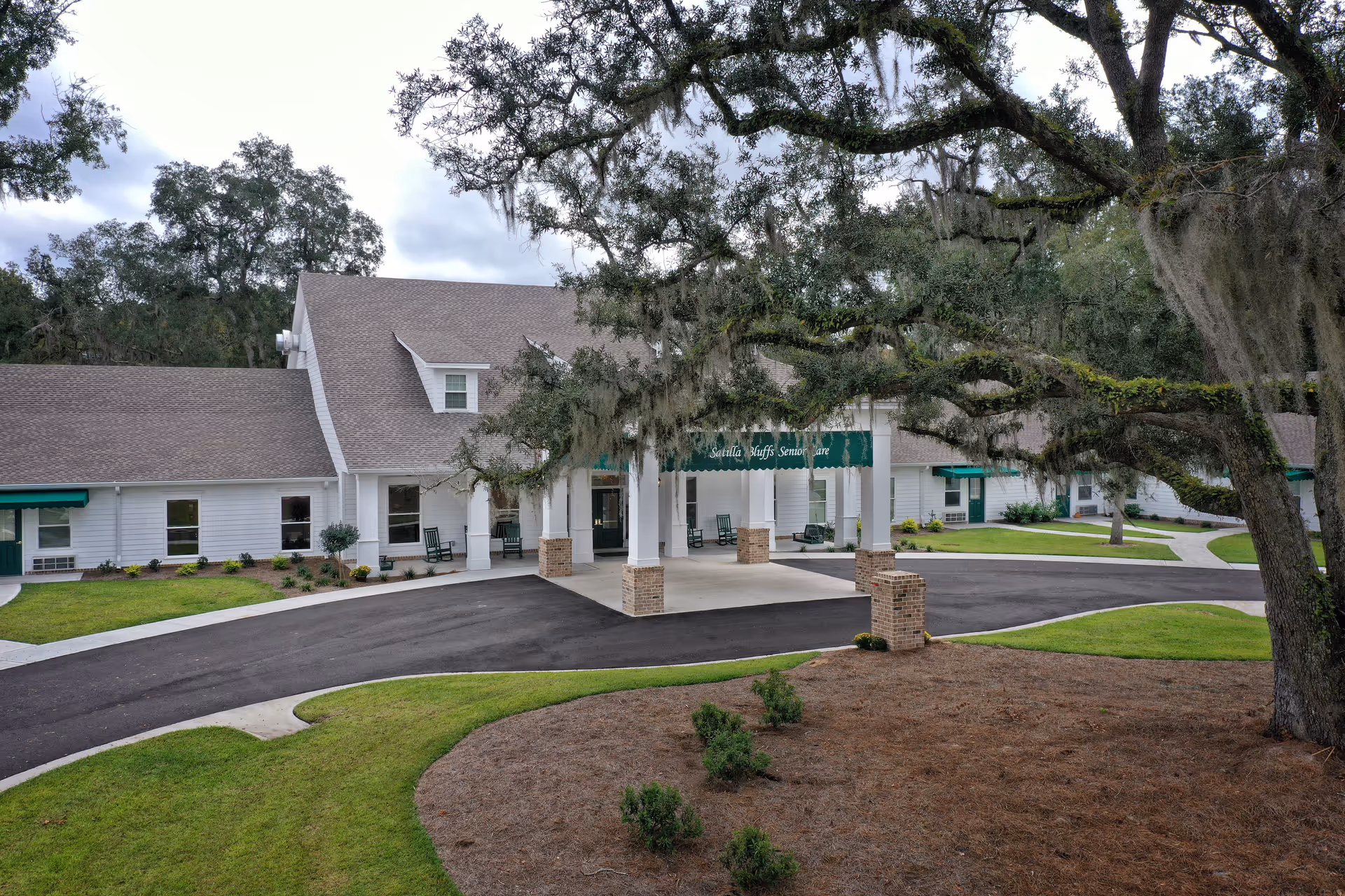 Front exterior of Satilla Bluffs Senior Care showing a covered entrance/portico, driveway, and a large oak tree in the foreground.