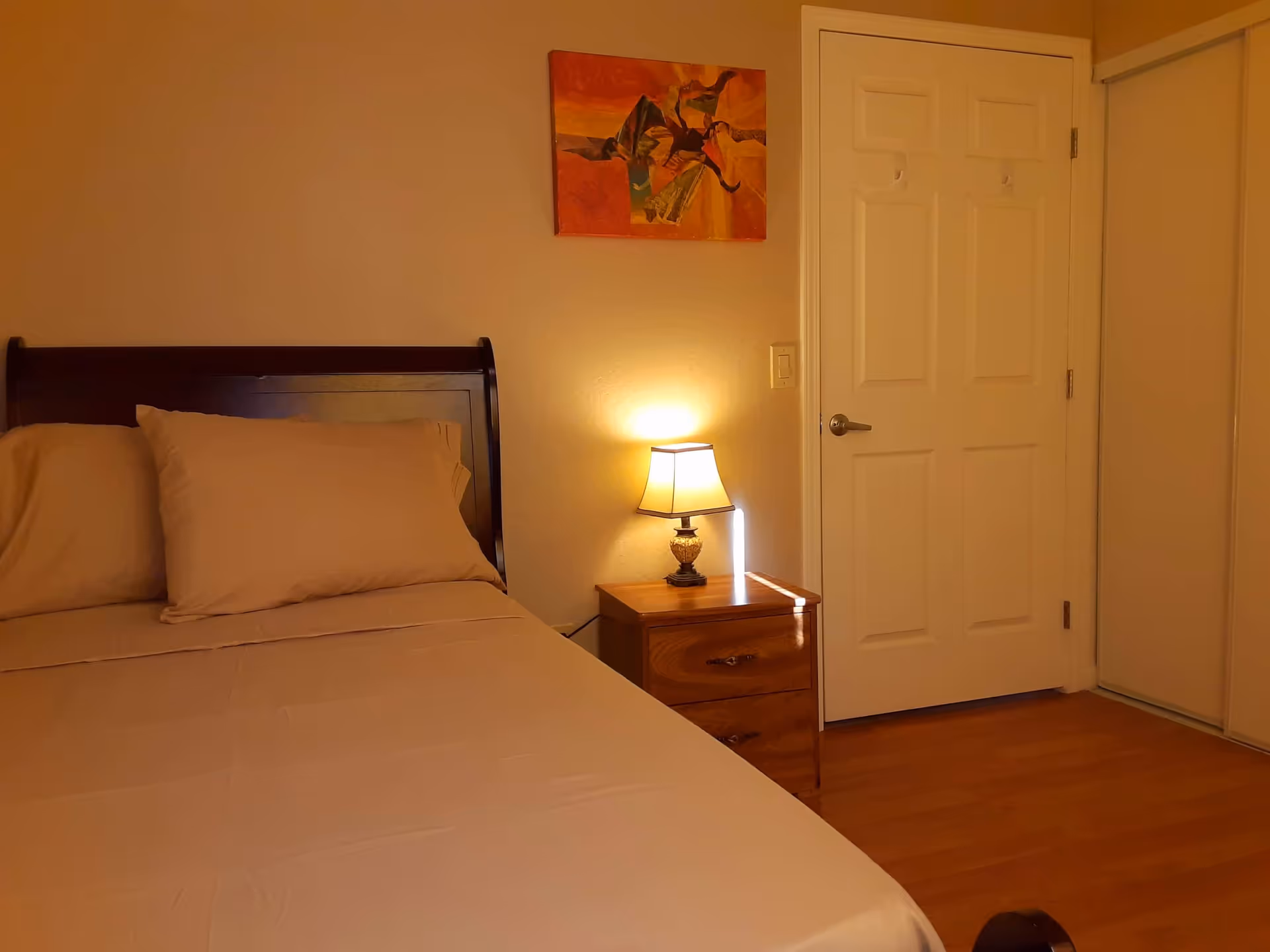 A tidy bedroom with a made bed, wooden headboard, nightstand holding a lit lamp, wall art, and a closed door.