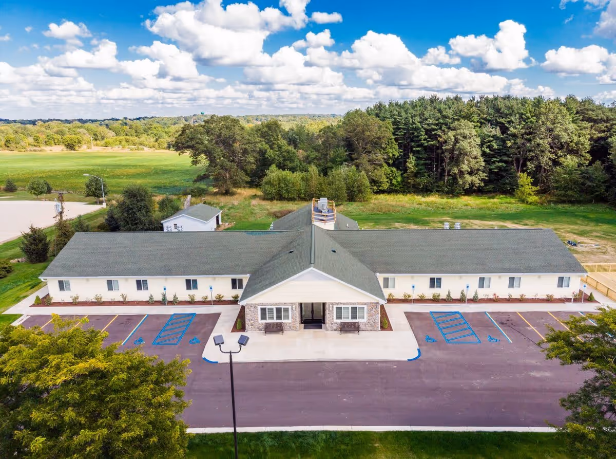 Aerial view of a single-story senior living building with a front entrance, parking lot with marked handicap spaces, and surrounding trees and fields.