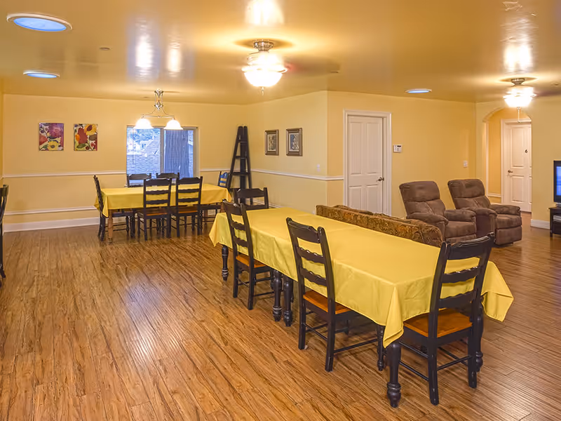 A spacious dining room in a senior living facility with two long tables covered with yellow tablecloths, surrounded by wooden chairs. The room has wood flooring, yellow walls, ceiling lights, and some wall decorations including paintings and framed pictures. In the background, there are two brown recliner chairs and a television.