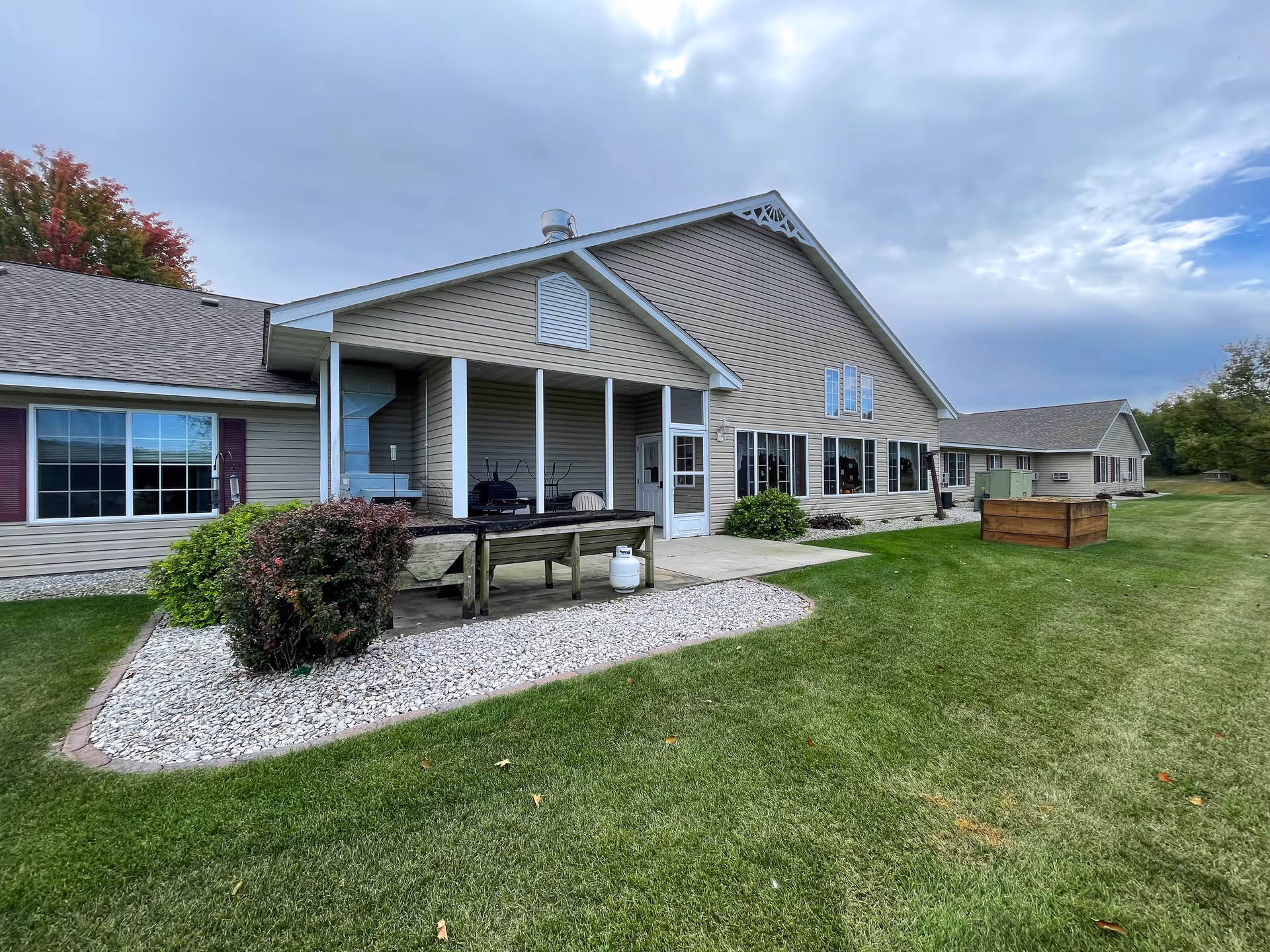 Beige single-story senior living building with a covered patio, large windows, and a landscaped lawn under a cloudy sky.
