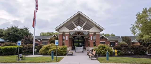 Front exterior view of Ben Atchley Tennessee State Veterans' Homes showing a brick building with a covered entrance supported by columns, benches along the walkway, two flagpoles with flags, and surrounding greenery under a cloudy sky.