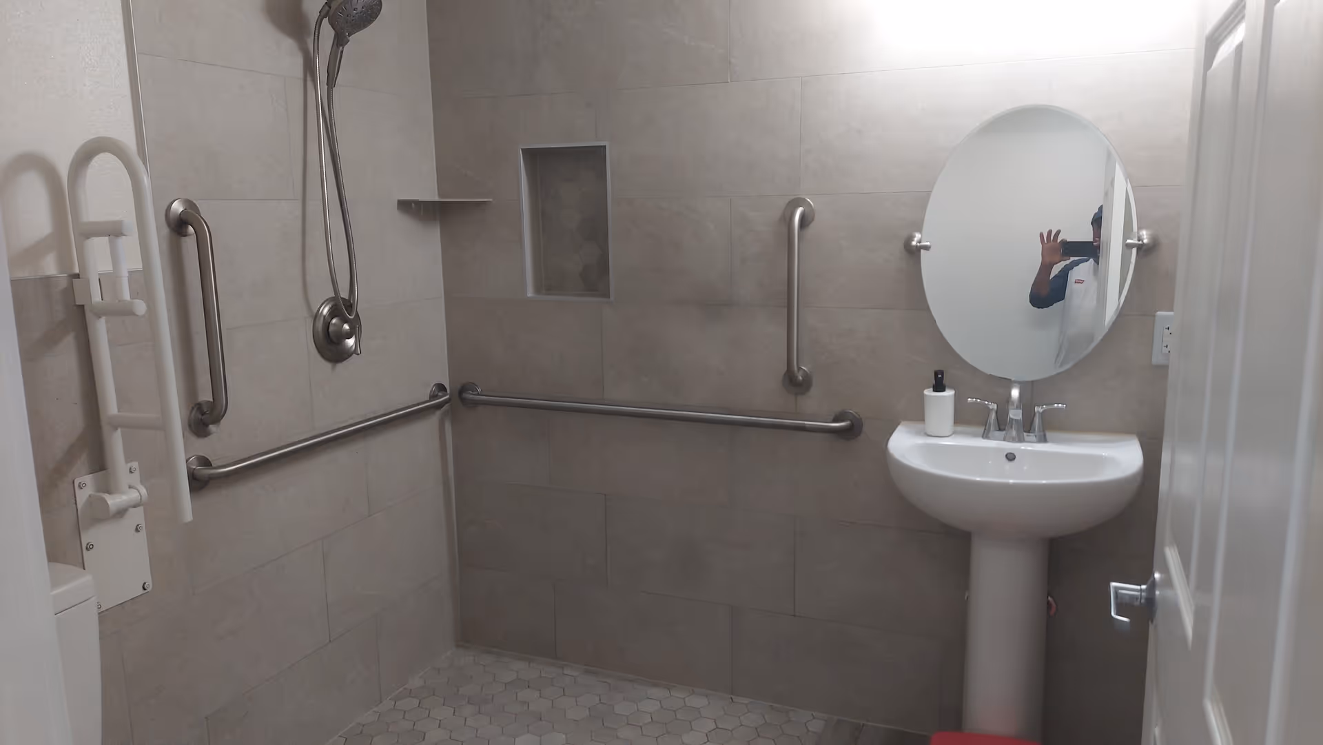Accessible bathroom with grab bars, a pedestal sink, handheld shower, and oval mirror.