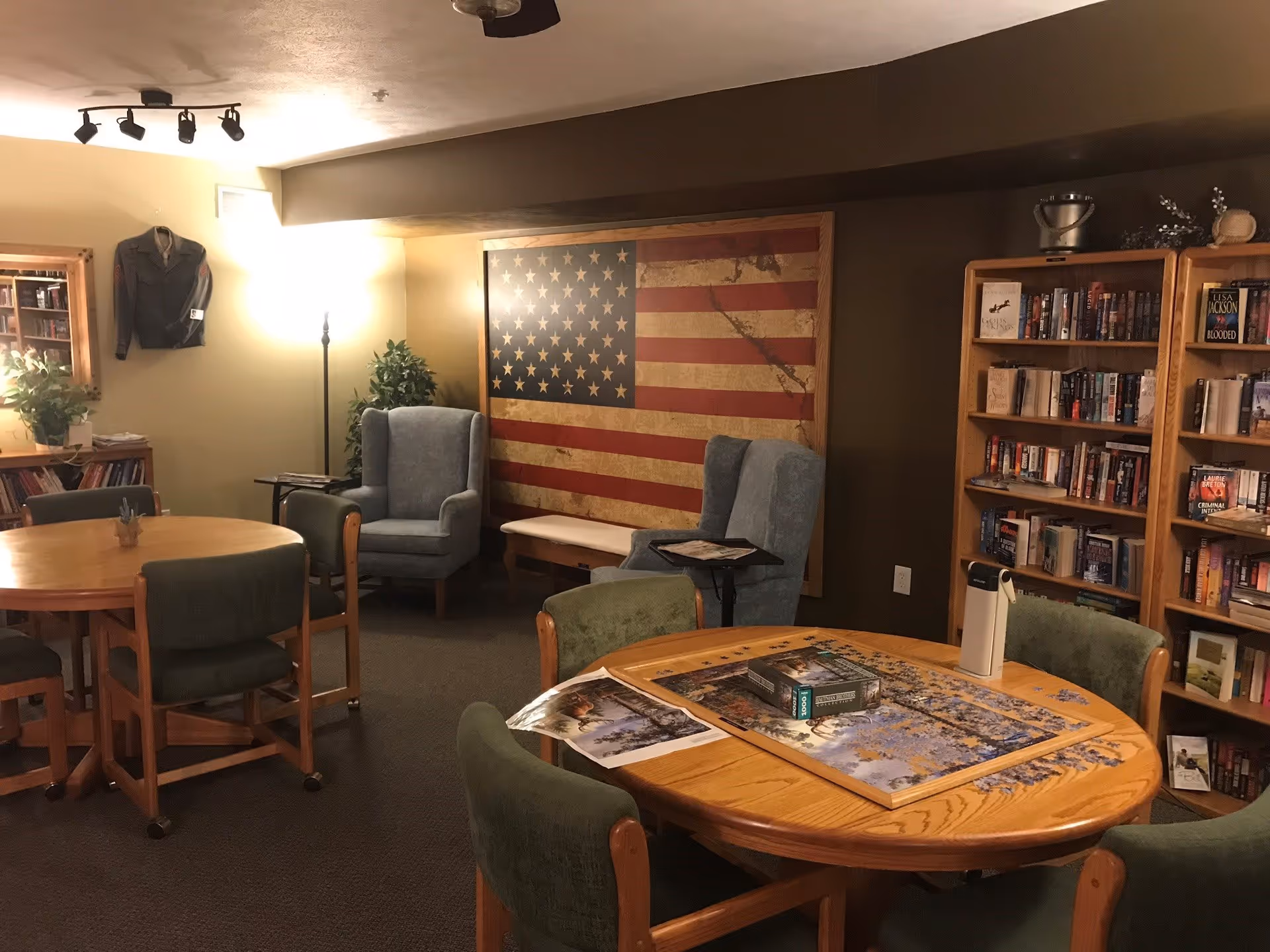 A cozy common room with round wooden tables and green cushioned chairs. One table has a partially completed jigsaw puzzle and a water bottle on it. There are two blue armchairs near a large framed American flag on the wall. Bookshelves filled with books line the right side of the room. A floor lamp and a framed jacket hanging on the wall add to the homey atmosphere.