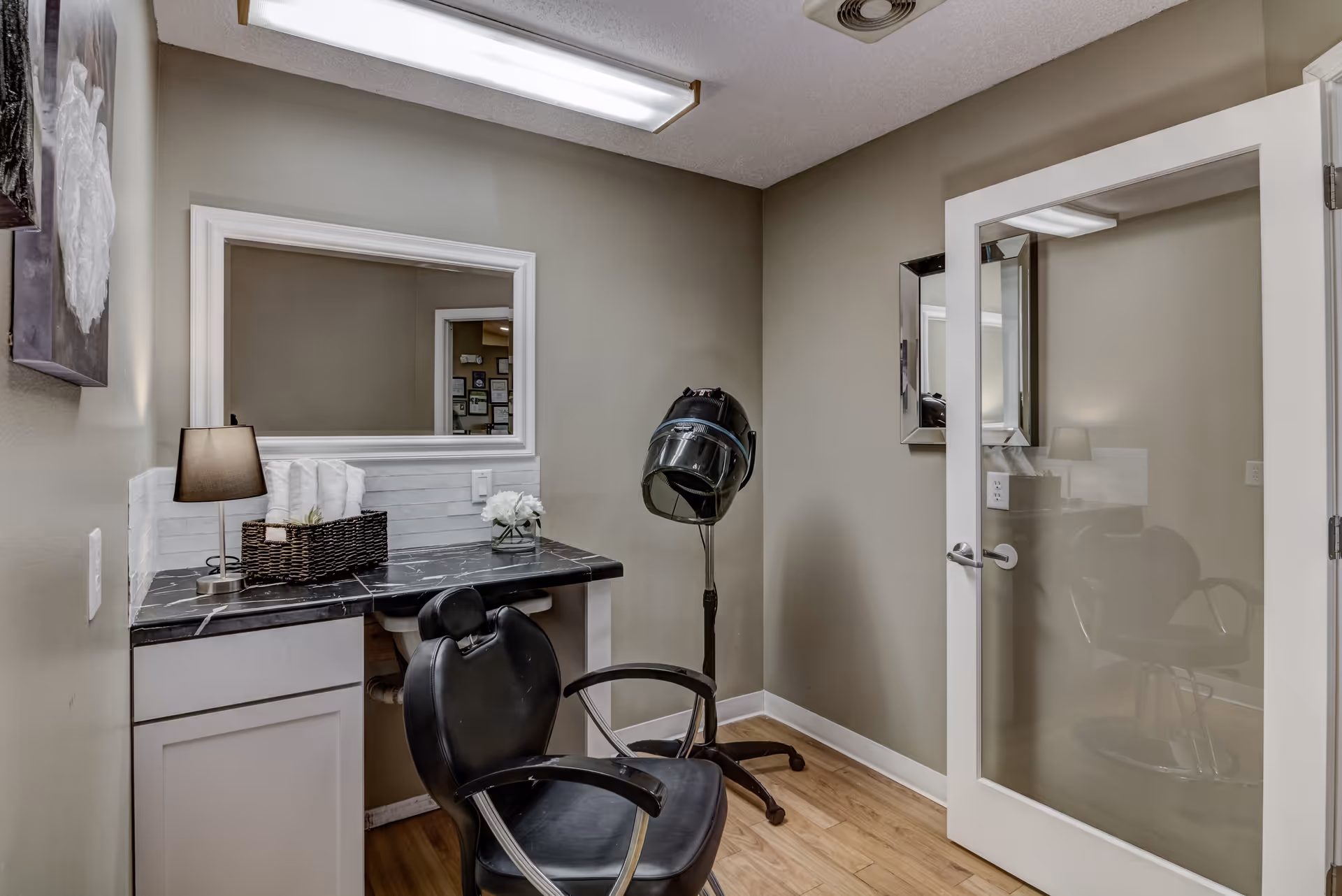 A small salon room with a black salon chair, a hair dryer hood on a stand, a black marble countertop with a lamp, a basket with towels, and a small flower arrangement. The room has beige walls, a large mirror above the countertop, and a glass door reflecting the interior.