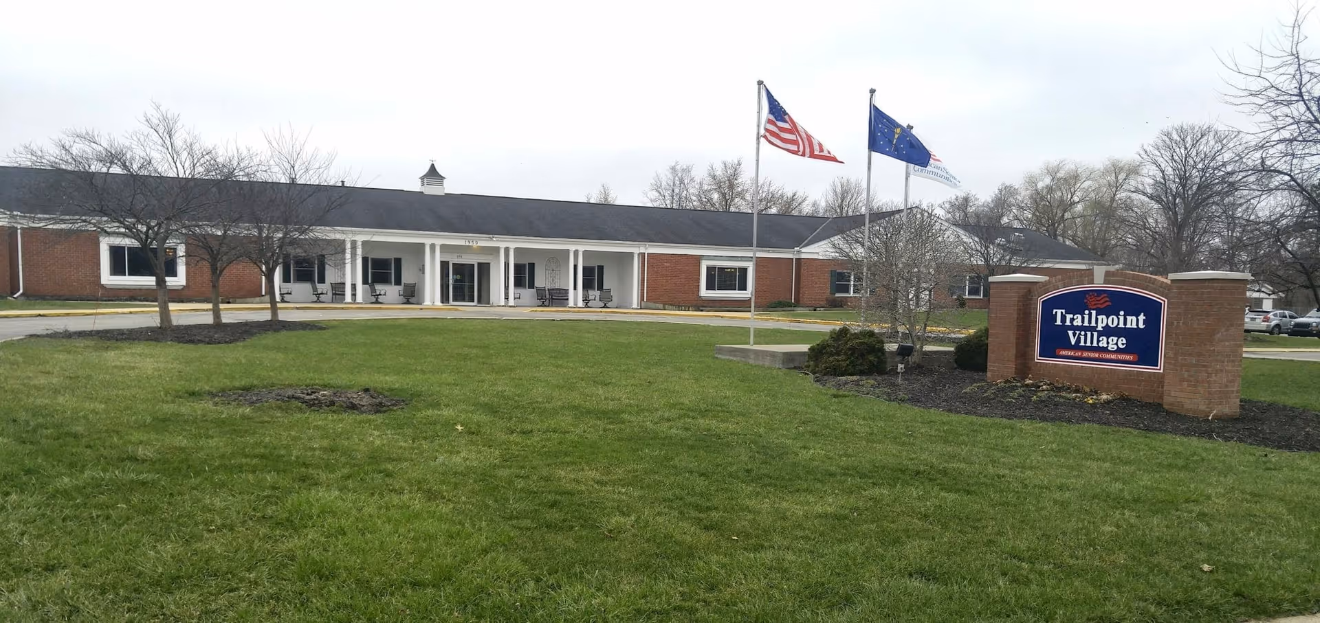 Single-story brick senior living facility with a covered entrance, flagpoles, and a 'Trailpoint Village' sign on a grassy lawn.