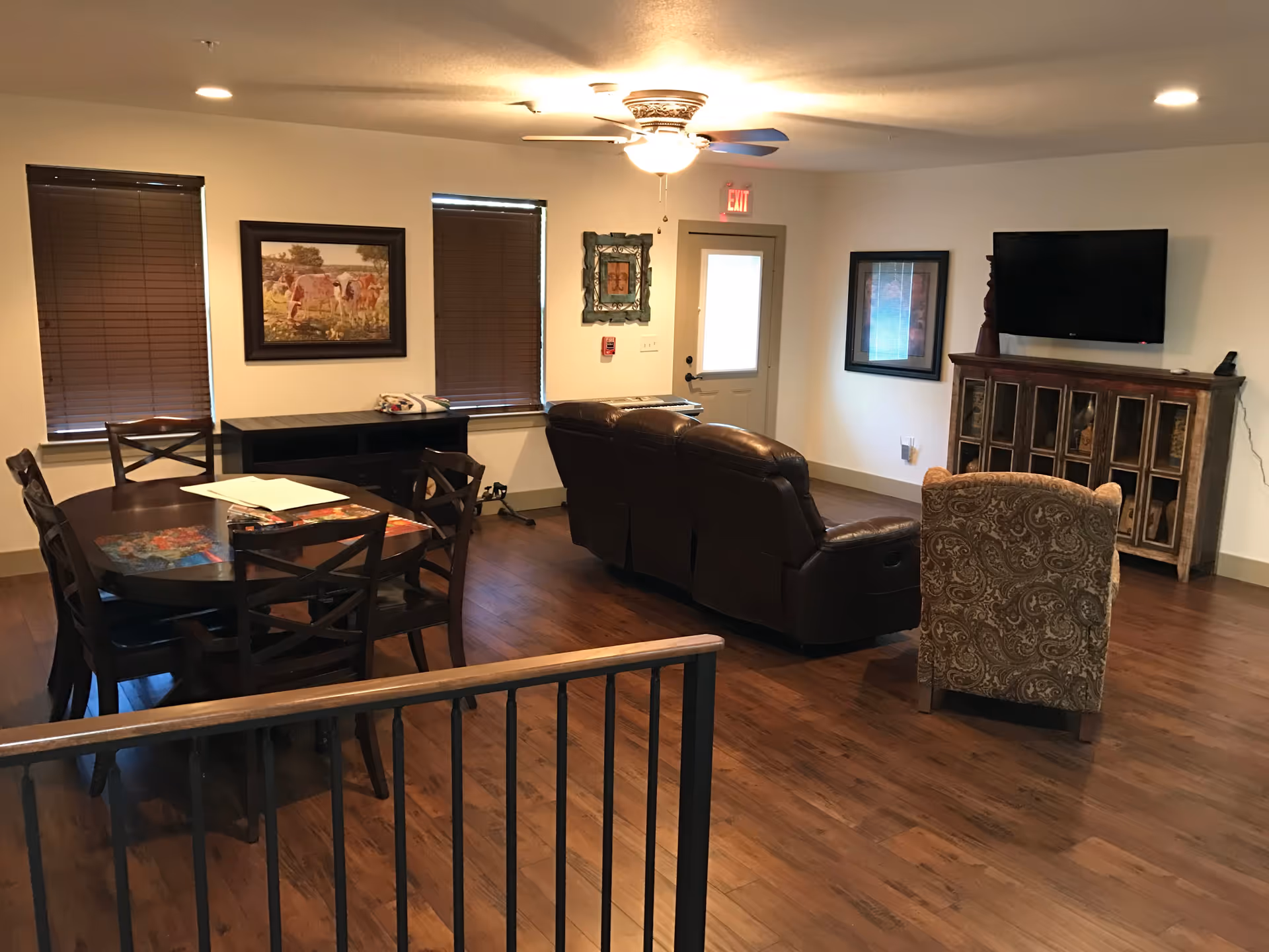 A spacious living room area with wooden flooring, featuring a dark wooden dining table with six chairs on the left, a brown leather sofa set in the center, and a patterned armchair facing a wall-mounted flat-screen TV above a rustic wooden cabinet. The room has two windows with closed blinds, framed artwork on the walls, a ceiling fan with lights, and a door with an exit sign above it.