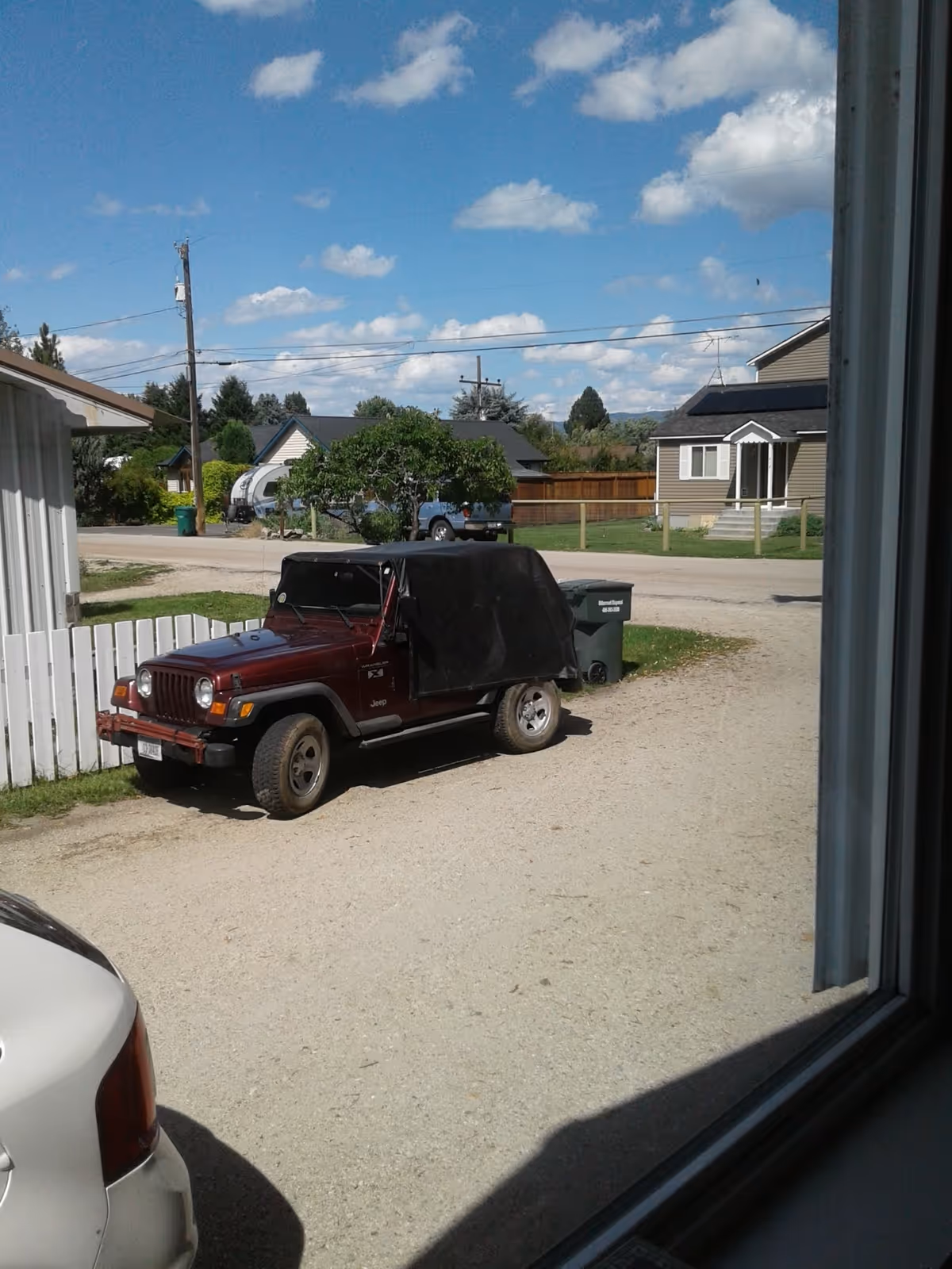 Red Jeep with a black soft top parked on a gravel driveway beside houses.