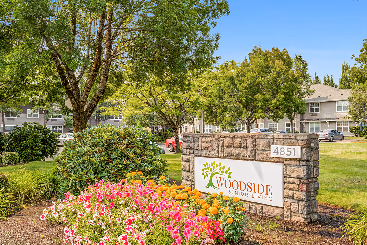 Stone sign reads Woodside Senior Living at the entrance, with colorful flowers and trees.