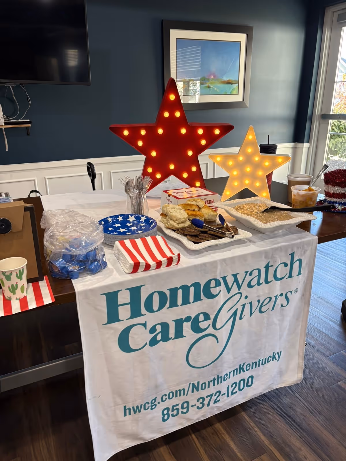 Indoor table setup with red/white star lights and Homewatch CareGivers banner.