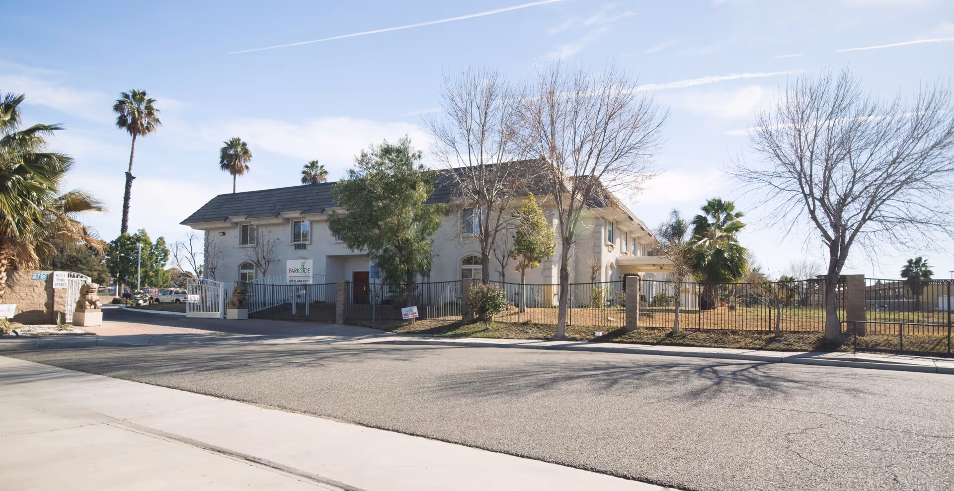 Exterior view of Parkside Mansion Assisted Living Community building with a fenced yard, several trees including palm trees, and a clear sky.
