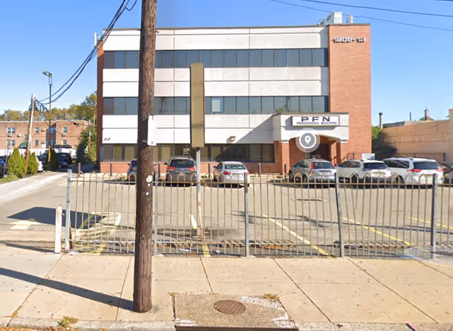 Front view of a three-story office building with a fenced parking lot and cars.