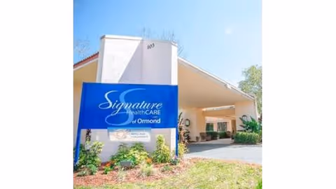 Exterior view of a healthcare facility with a large blue sign reading 'Signature HealthCARE of Ormond' in front of a white building with a covered entrance and some greenery around.