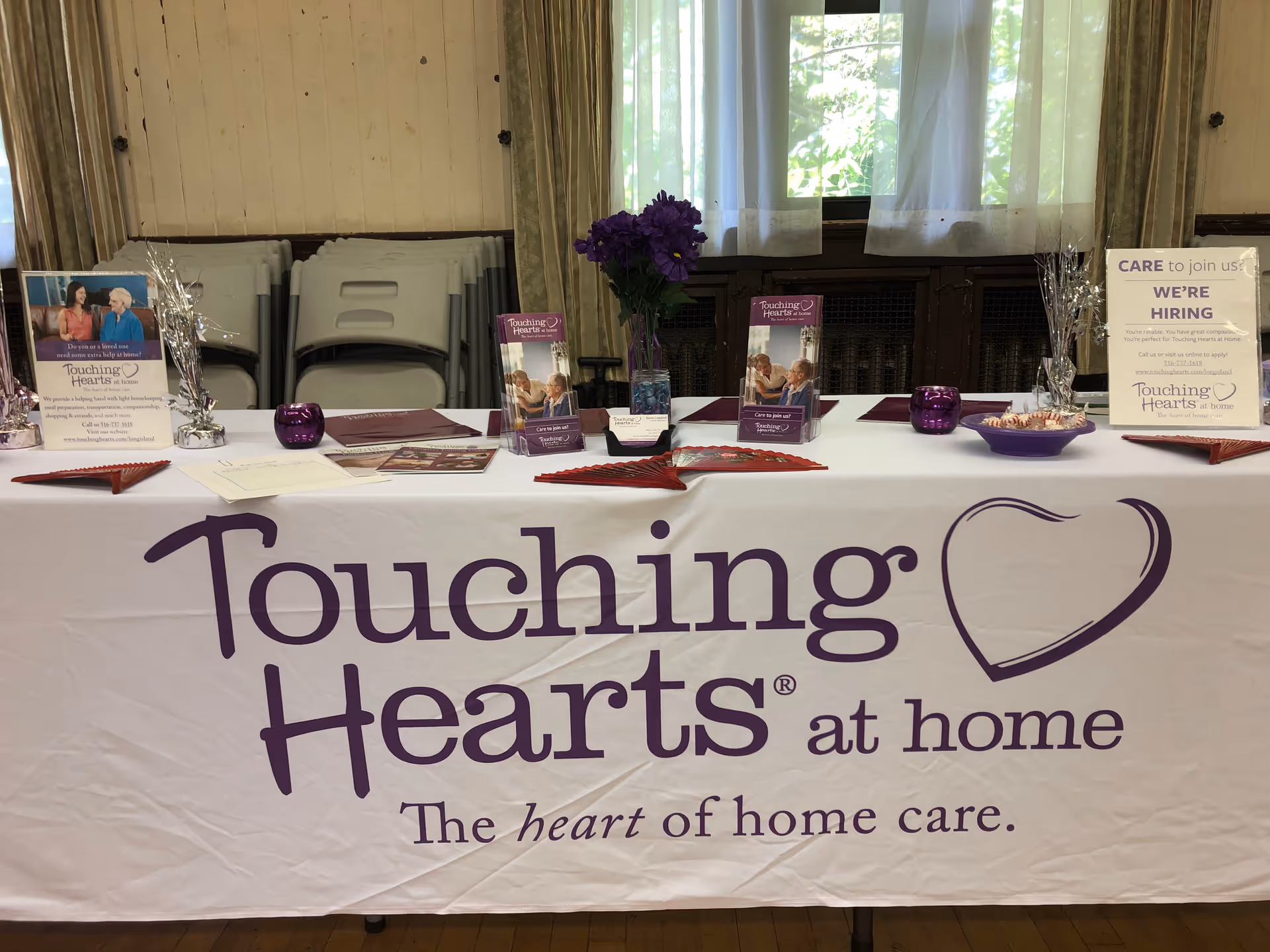Indoor event table draped with Touching Hearts at home banner, brochures, and purple decor.