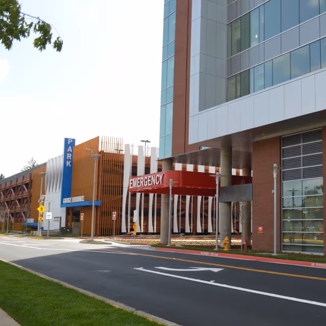 Exterior view of a modern hospital entrance labeled Emergency, with parking and glass facade.