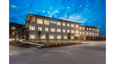 Exterior view of a three-story senior living facility named The Reserve at Lake Austin during twilight, with lights on inside the building and a mostly empty parking lot in the foreground.