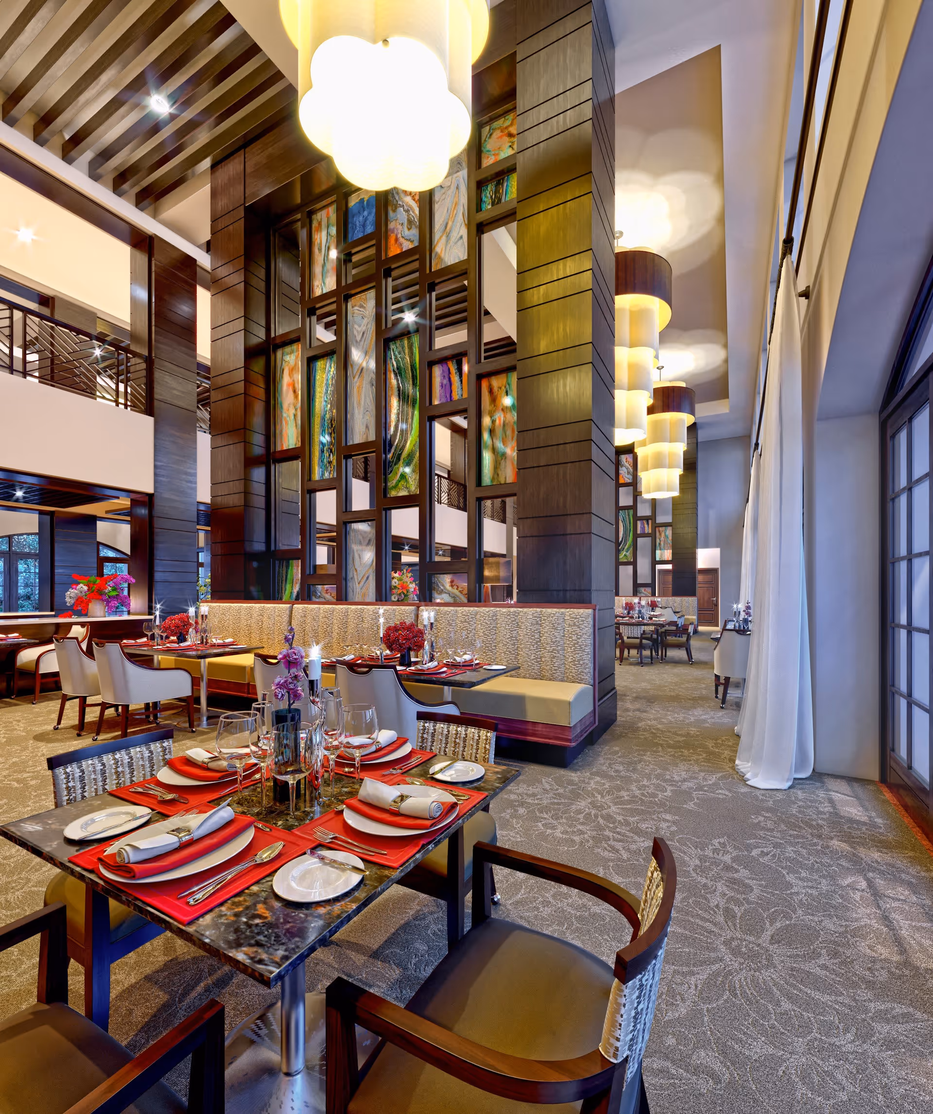 Elegant dining room with tables set for a meal, featuring red placemats, white napkins, and glassware. The room has high ceilings with large pendant lights, decorative wooden columns with colorful abstract panels, and large windows with white curtains allowing natural light.