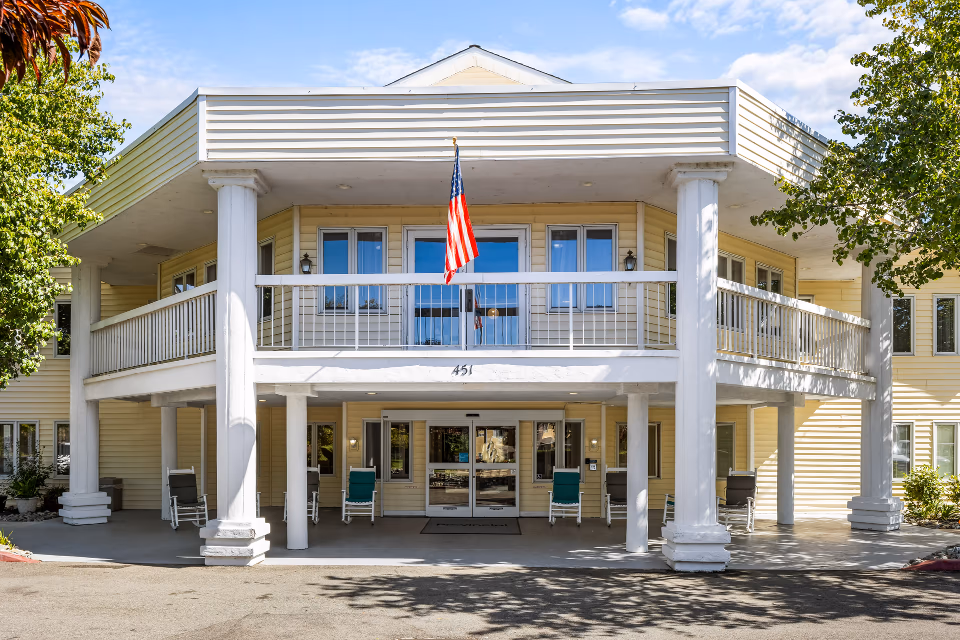Front exterior view of a two-story senior living facility building with a covered entrance supported by white columns. There are rocking chairs on the porch, an American flag hanging above the entrance, and trees partially framing the building under a clear blue sky.
