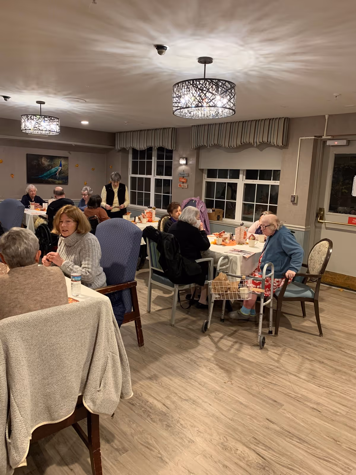 A group of elderly residents seated at tables in a communal dining room of a senior living facility.