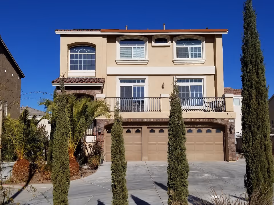 Two-story tan senior living home with garage doors, tall cypress trees, and blue sky.