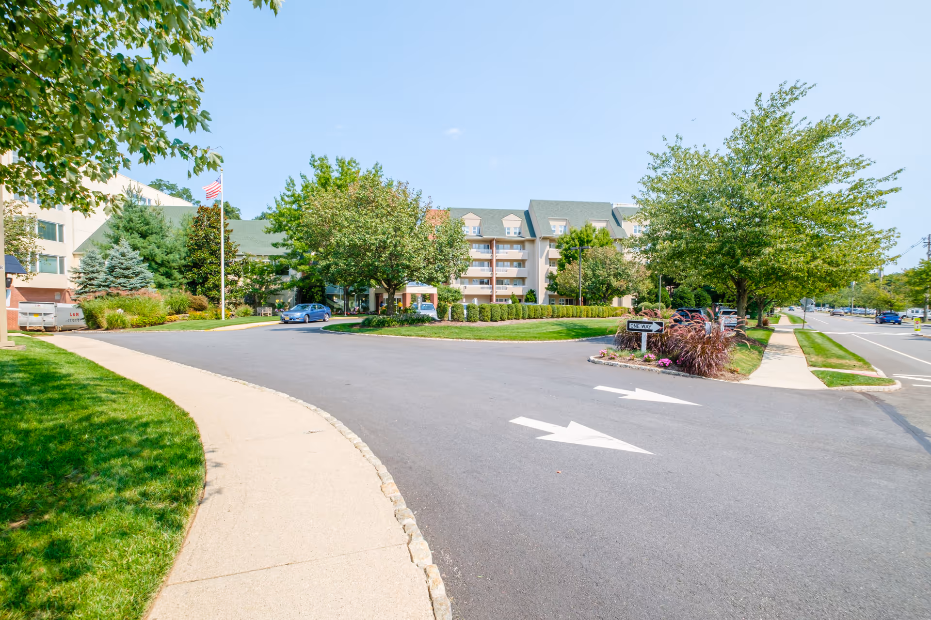 Driveway and landscaped circular entrance leading to the front of a multi-story senior living building with trees and an American flag.