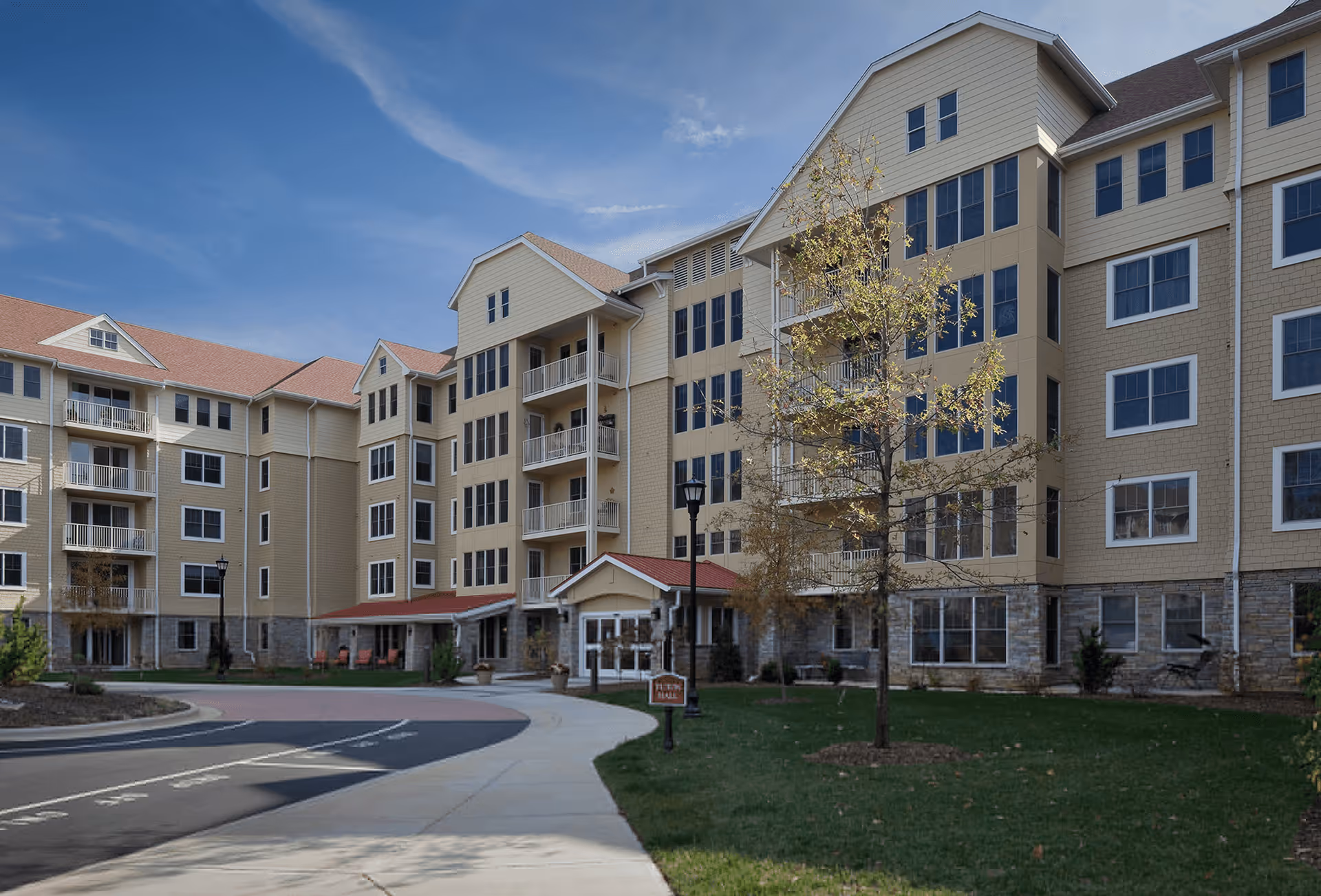 Front exterior of a multi-story beige senior living building with balconies, an entrance canopy, driveway, and landscaped lawn.