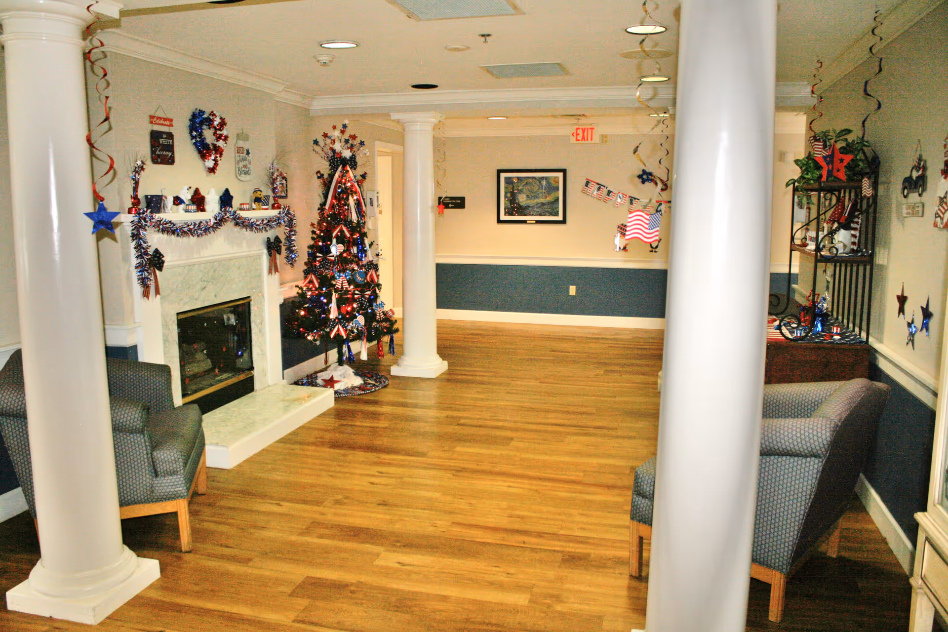 Interior common area decorated with patriotic red, white, and blue decorations including a Christmas tree, garlands, and hanging stars. The room features wooden flooring, white columns, a white fireplace, and two upholstered chairs on either side.
