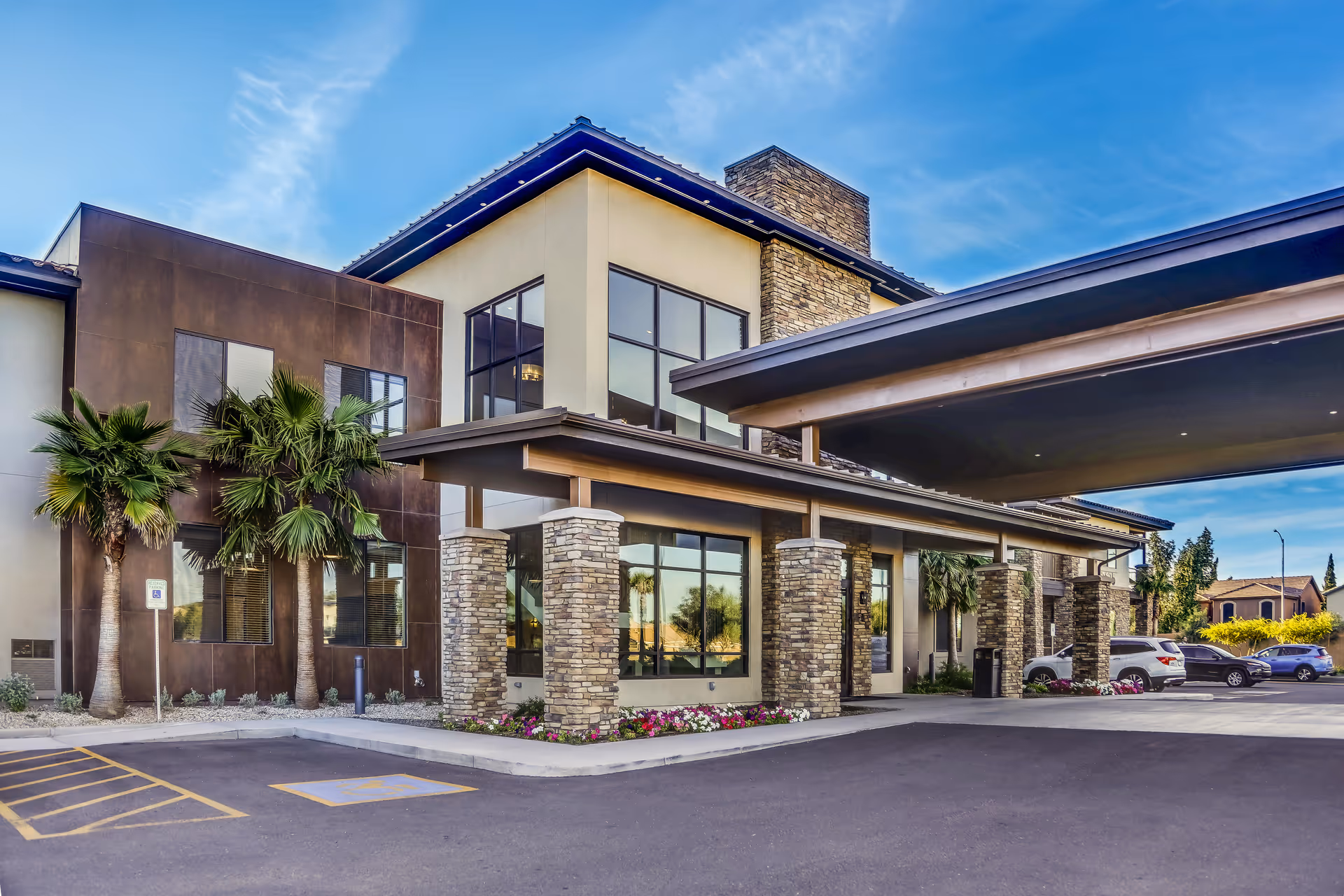 Covered entrance and facade of a modern senior living facility with stone pillars, large windows, palm trees, and parked cars.