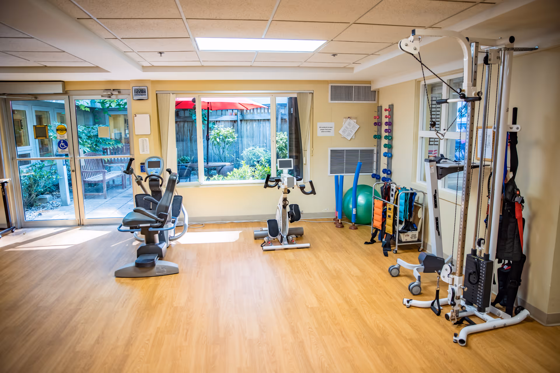 A bright exercise room with wooden flooring featuring stationary exercise bikes, a weight machine, dumbbells, exercise balls, and resistance bands. Large windows and glass doors provide a view of an outdoor garden area with a red umbrella and greenery.