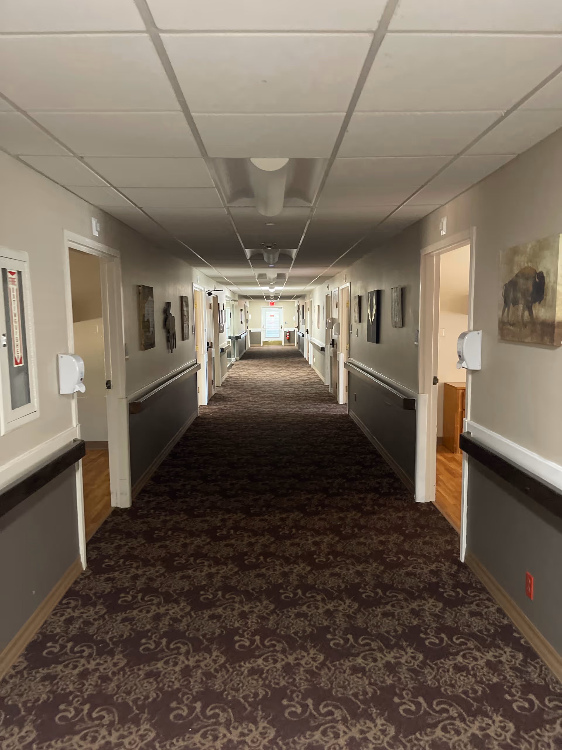 Long, well-lit hallway in a senior living facility with doors and wall art.