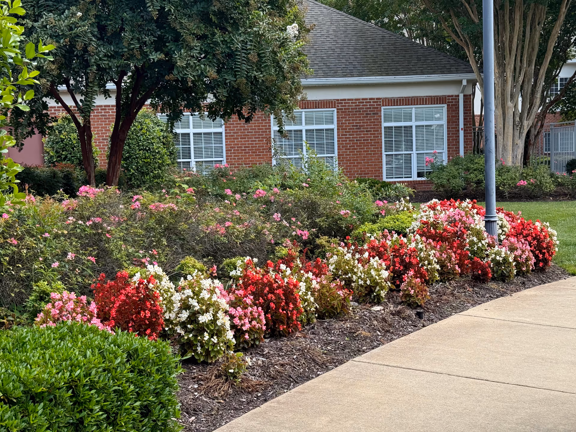 Brick building front with windows, lush landscaping, and a lamppost along a sidewalk.