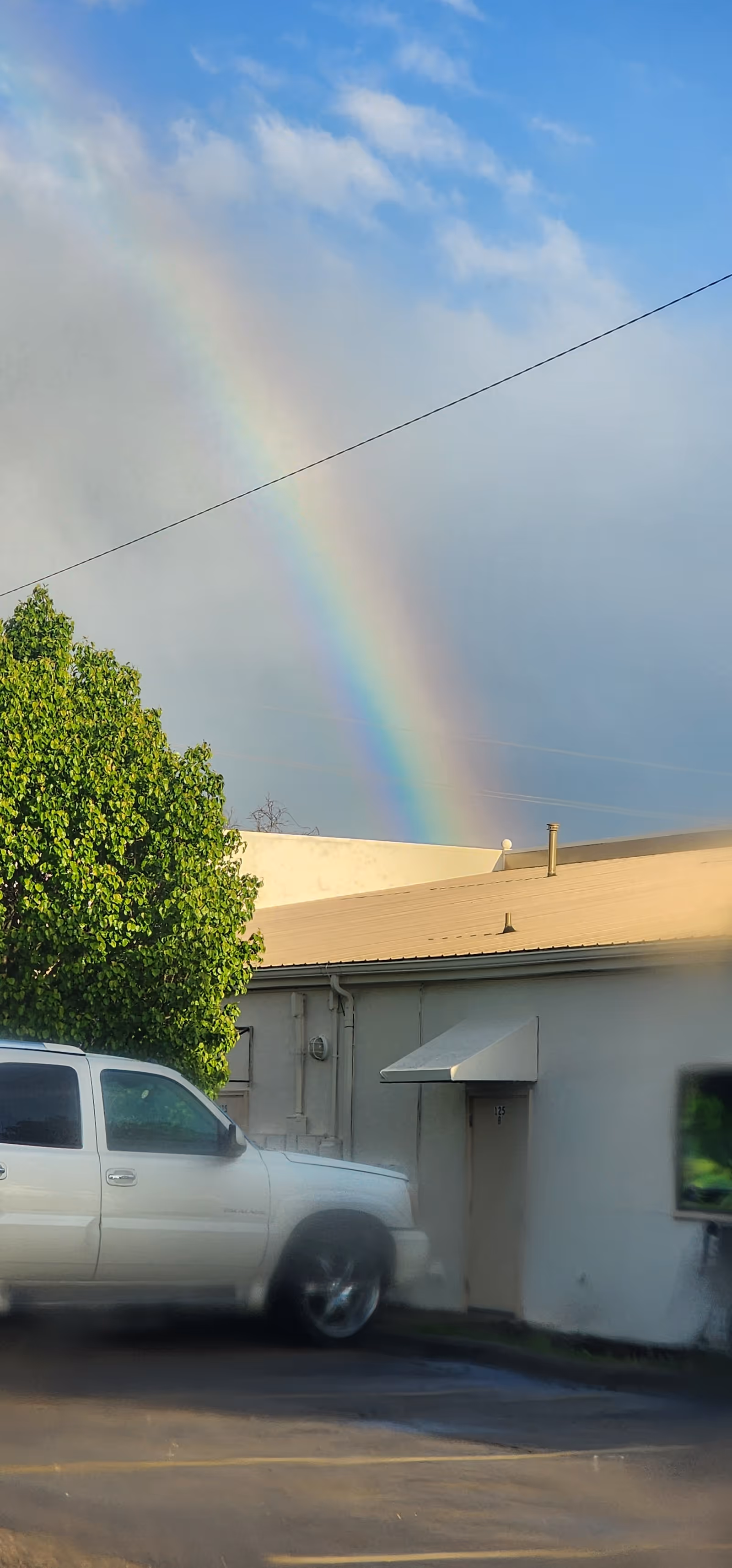 Rainbow arches over a white building with a tree and a parked white truck.