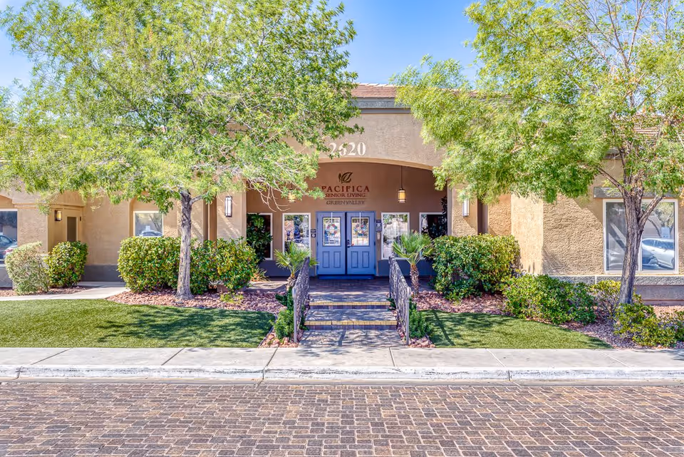 Front entrance of a senior living building with double doors, a small ramp and steps, and landscaped trees and shrubs.