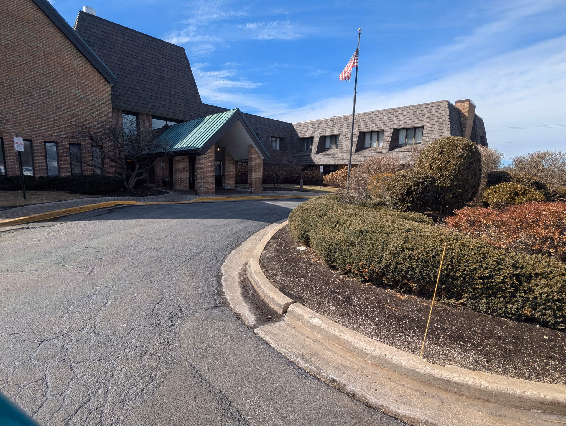 Exterior of a senior living facility with brick building, driveway, shrubs, and flag.