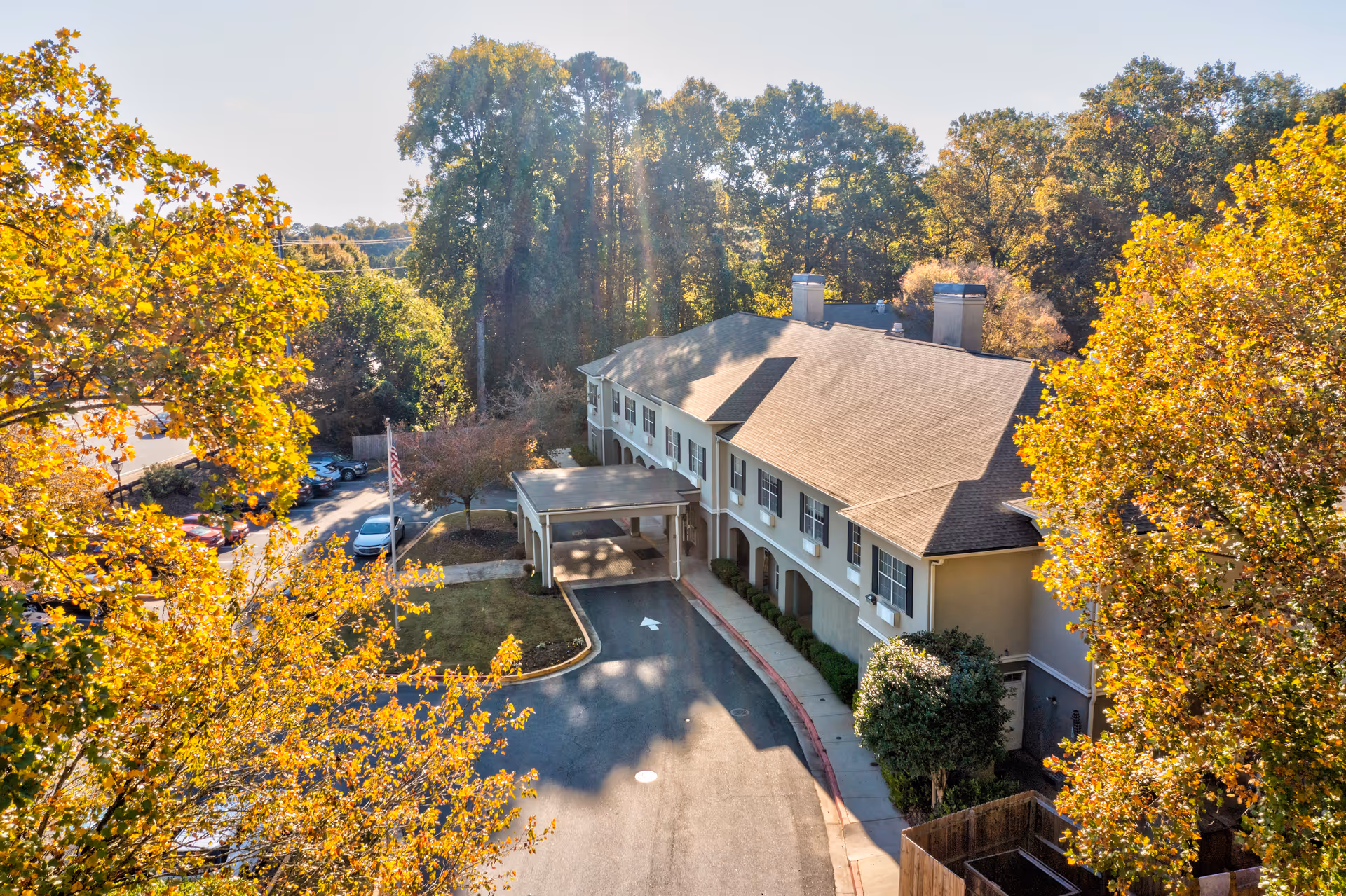 Aerial view of a two-story assisted living facility building surrounded by trees with autumn foliage. The building has a covered entrance driveway and a parking area with several cars. The scene is bathed in sunlight with a clear sky.