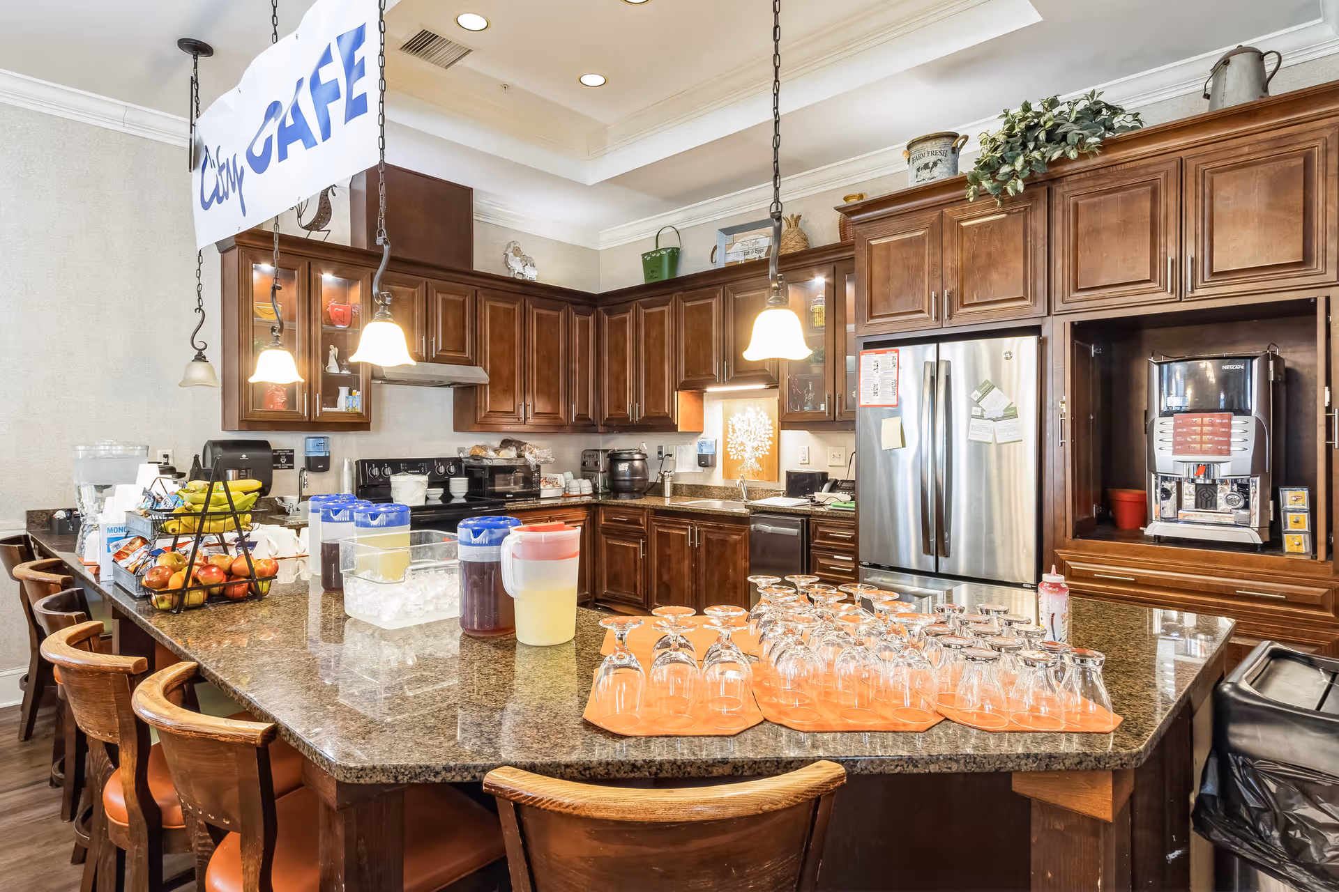 Interior view of a kitchen area in a senior living facility with a large granite countertop island surrounded by wooden chairs. The countertop holds several pitchers of drinks, a container of ice, a basket of fruit, and many upside-down glasses on orange mats. The kitchen has dark wooden cabinets, a stainless steel refrigerator, a coffee machine, and hanging pendant lights. A sign hanging from the ceiling reads 'City CAFE'.