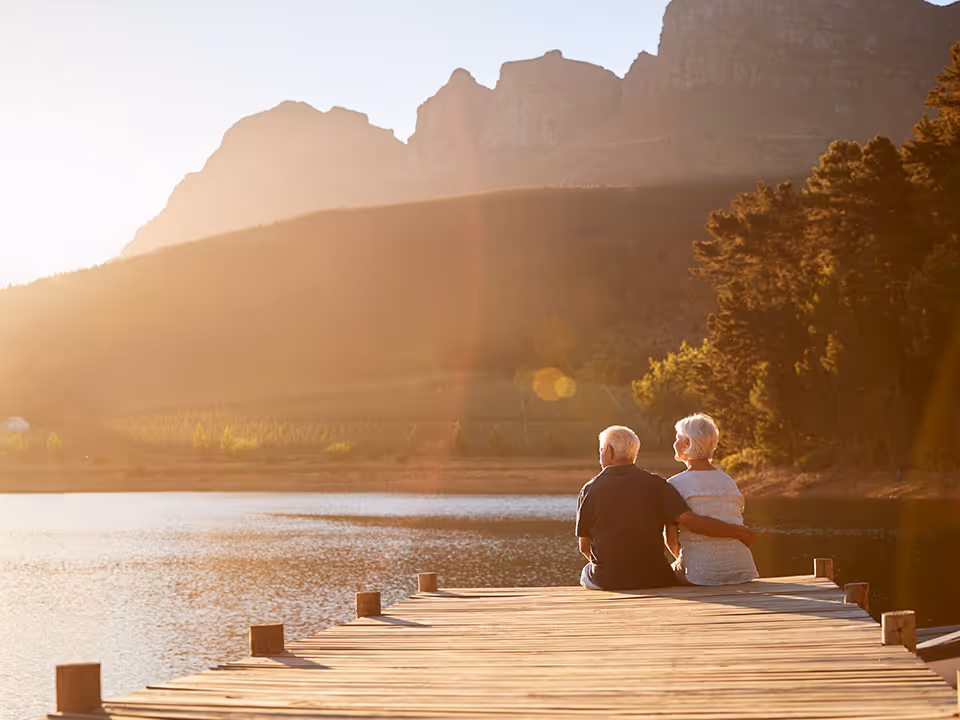 Two seniors sit on a wooden dock overlooking a lake at sunset.