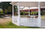 White gazebo with lattice fence in a grassy courtyard, benches nearby, brick building in background.