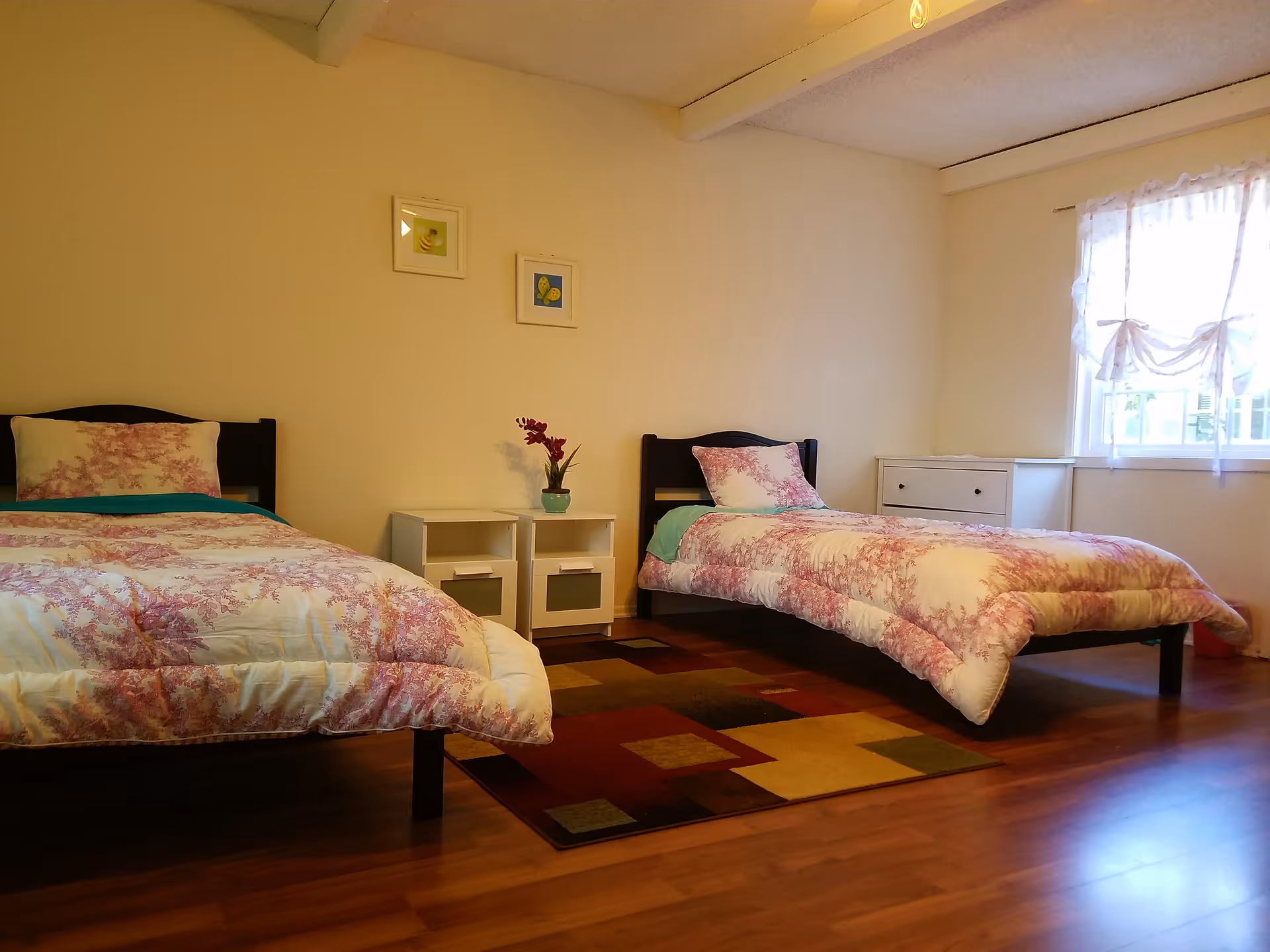 Bright twin bedroom with two single beds, floral bedding, a nightstand, rug, and a window with sheer curtains.