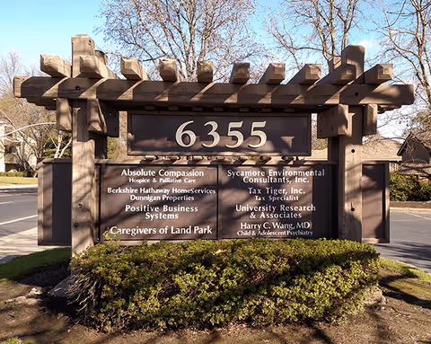 Large wooden sign at entrance lists Caregivers of Land Park LLC outdoors.