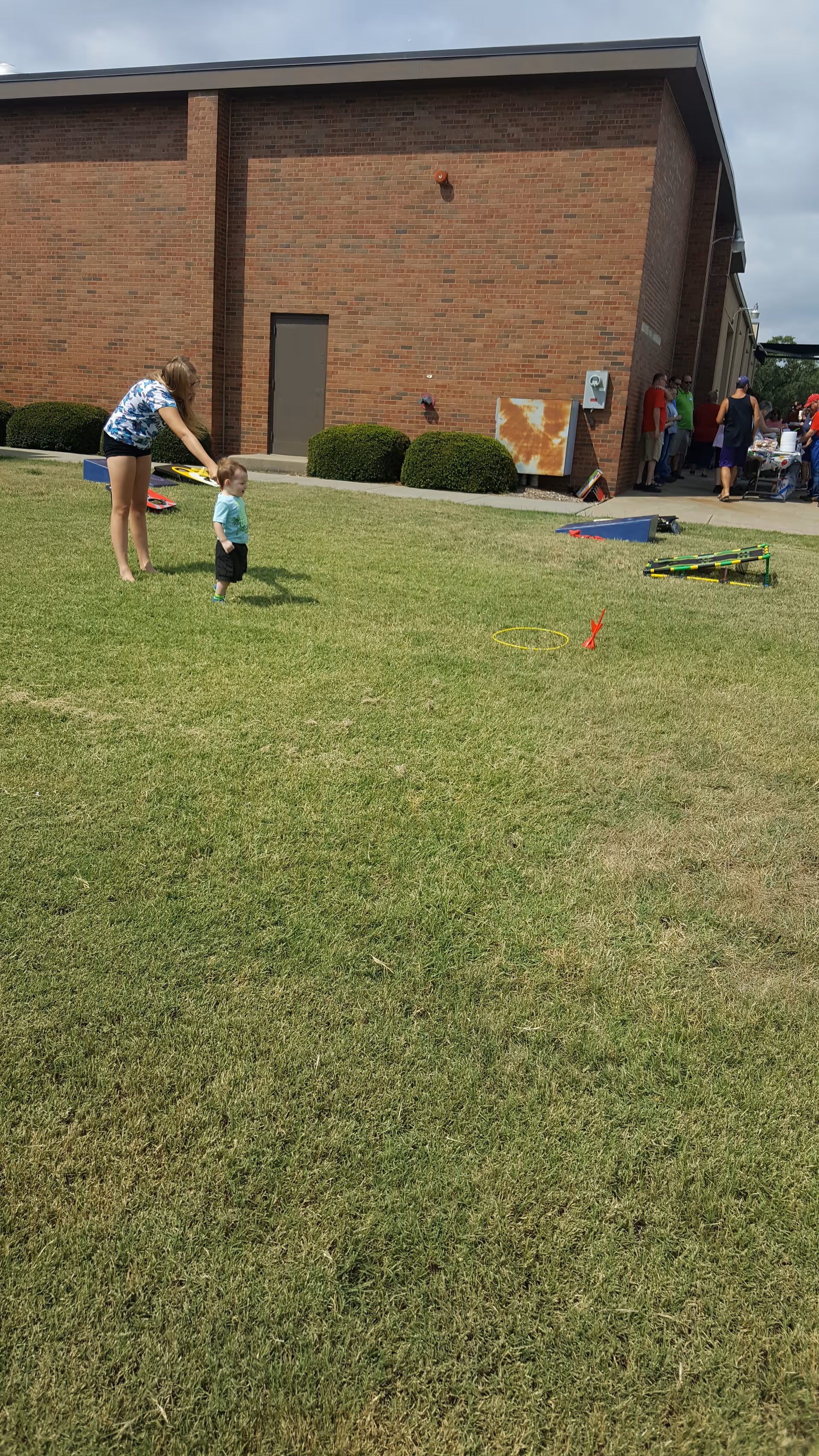 Adult guides a toddler on a grassy outdoor area beside a brick building.