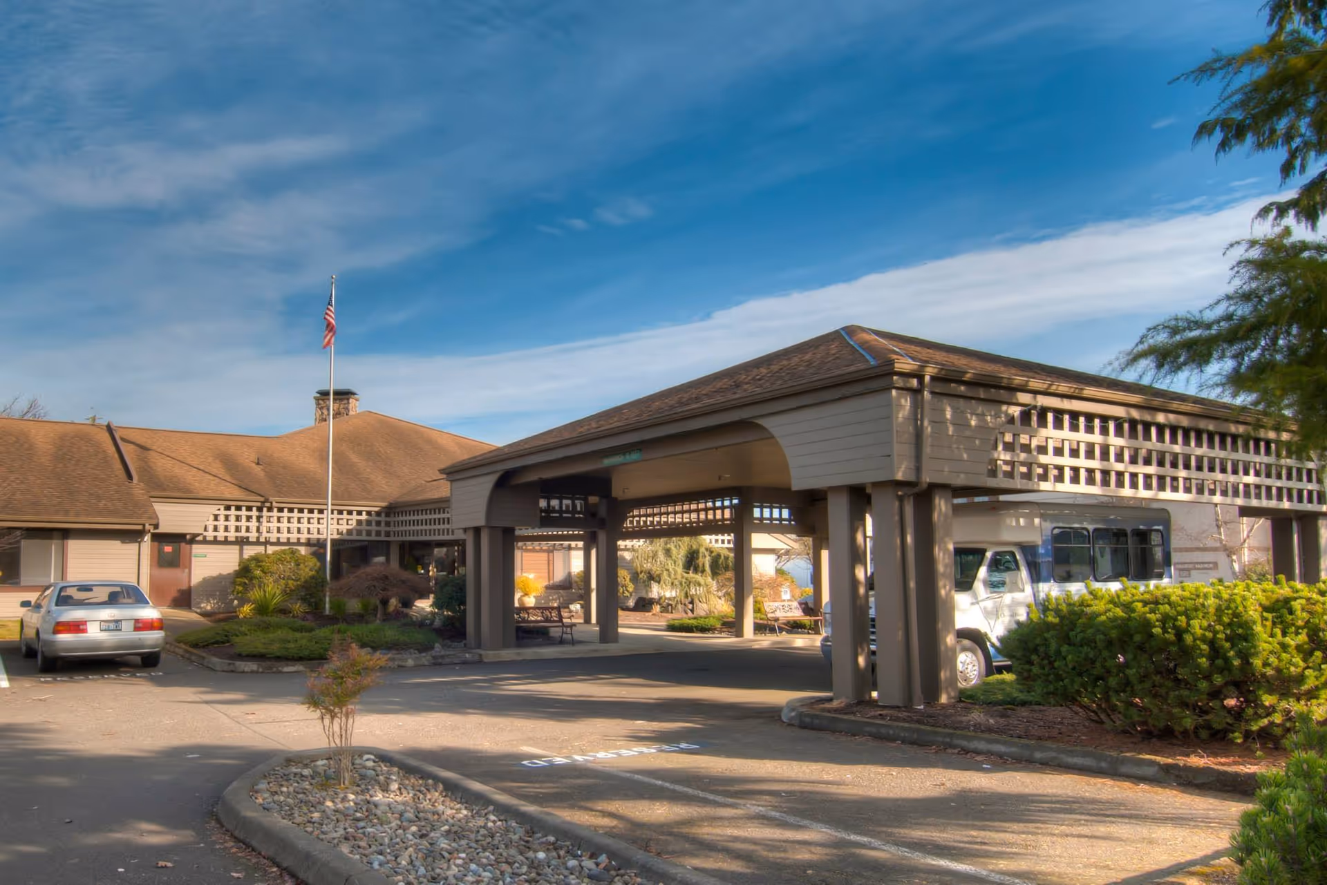 Exterior view of Channel Point Village senior living facility showing the main entrance with a covered drop-off area, an American flag on a flagpole, a parked car, a shuttle bus, and landscaped greenery under a blue sky with some clouds.