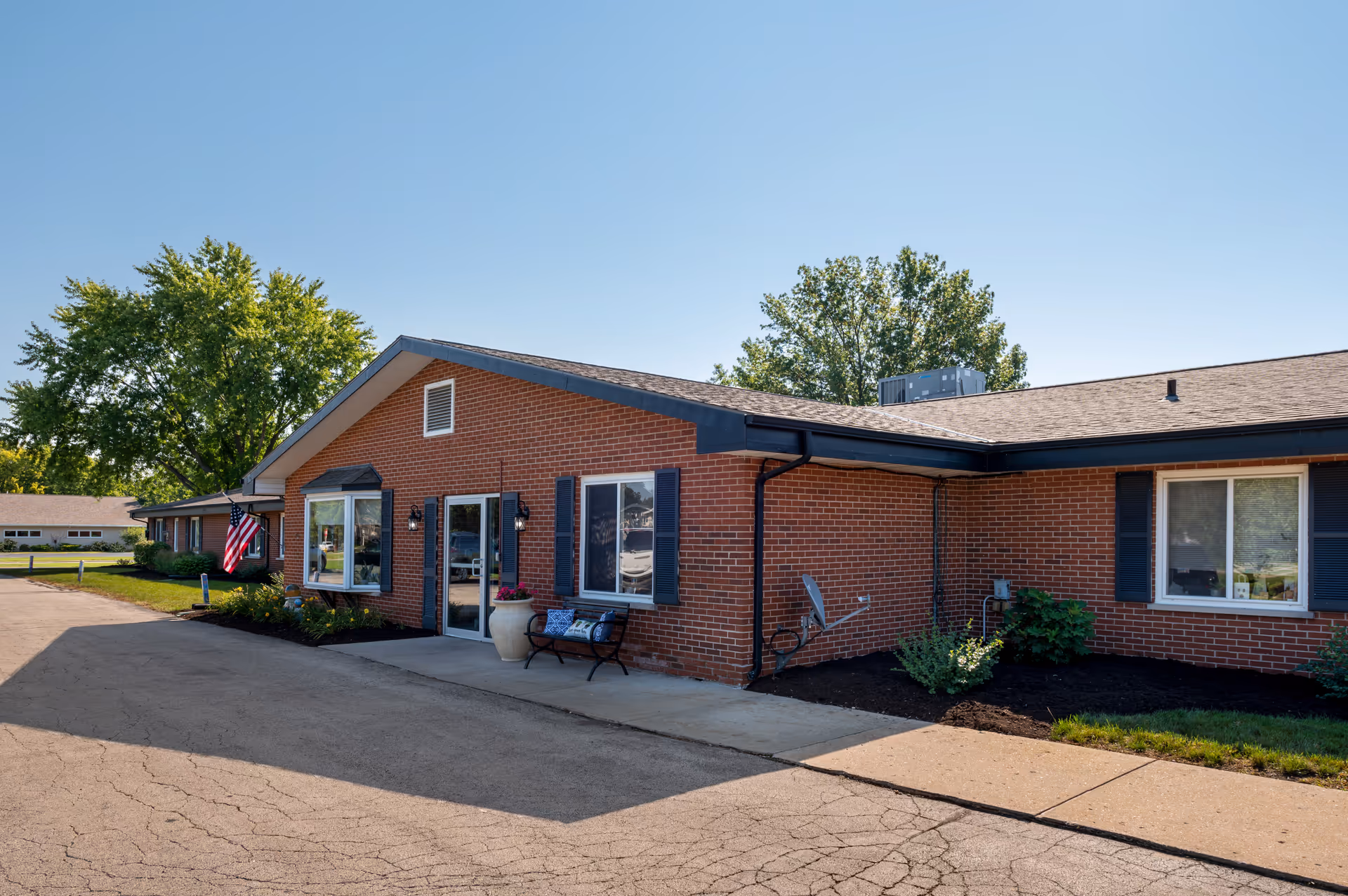 Red brick senior living facility exterior with entrance, windows, flag, and benches.