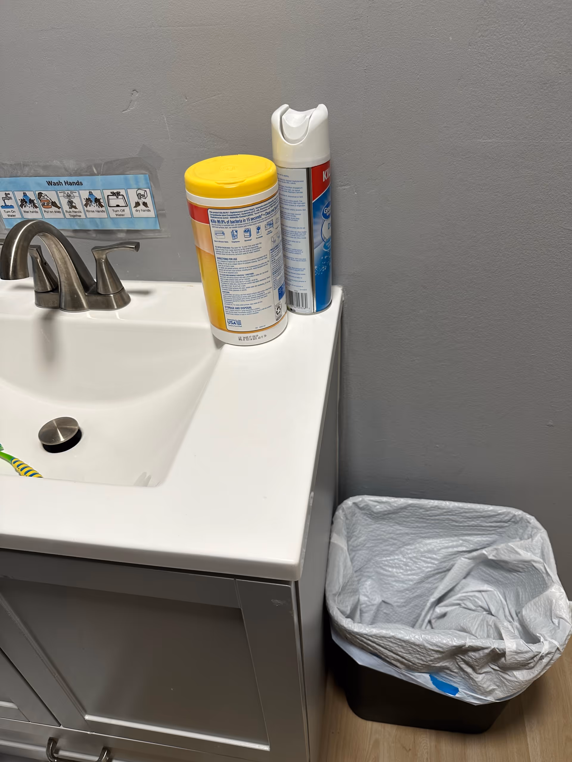 White bathroom sink with sanitizer container, wipes, spray can, and trash bin against gray wall.